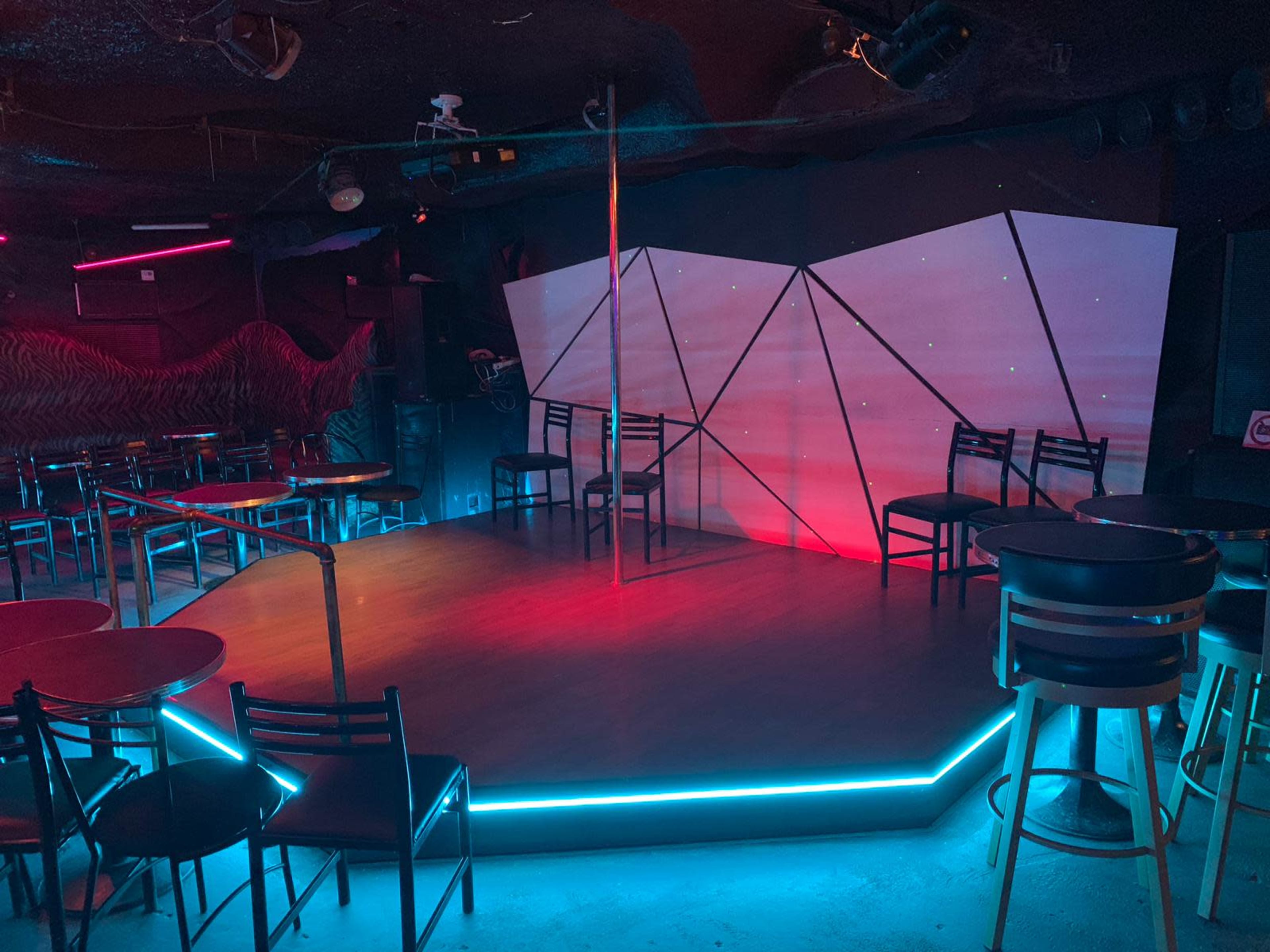 The image shows a dimly lit club interior featuring a stage with a pole, surrounded by circular tables and chairs.
