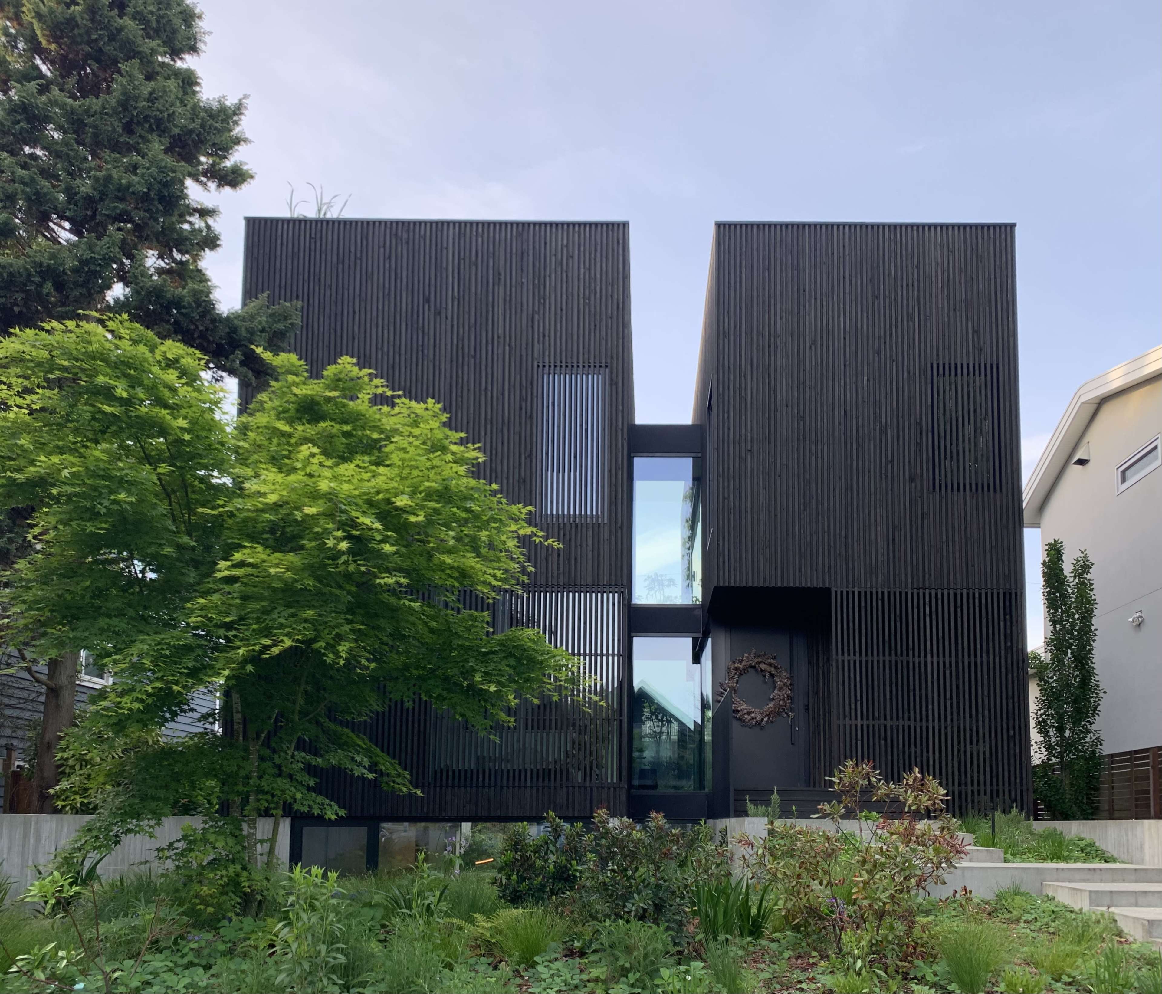 A modern black wooden house with two distinct rectangular sections, flanked by greenery and a landscaped garden.