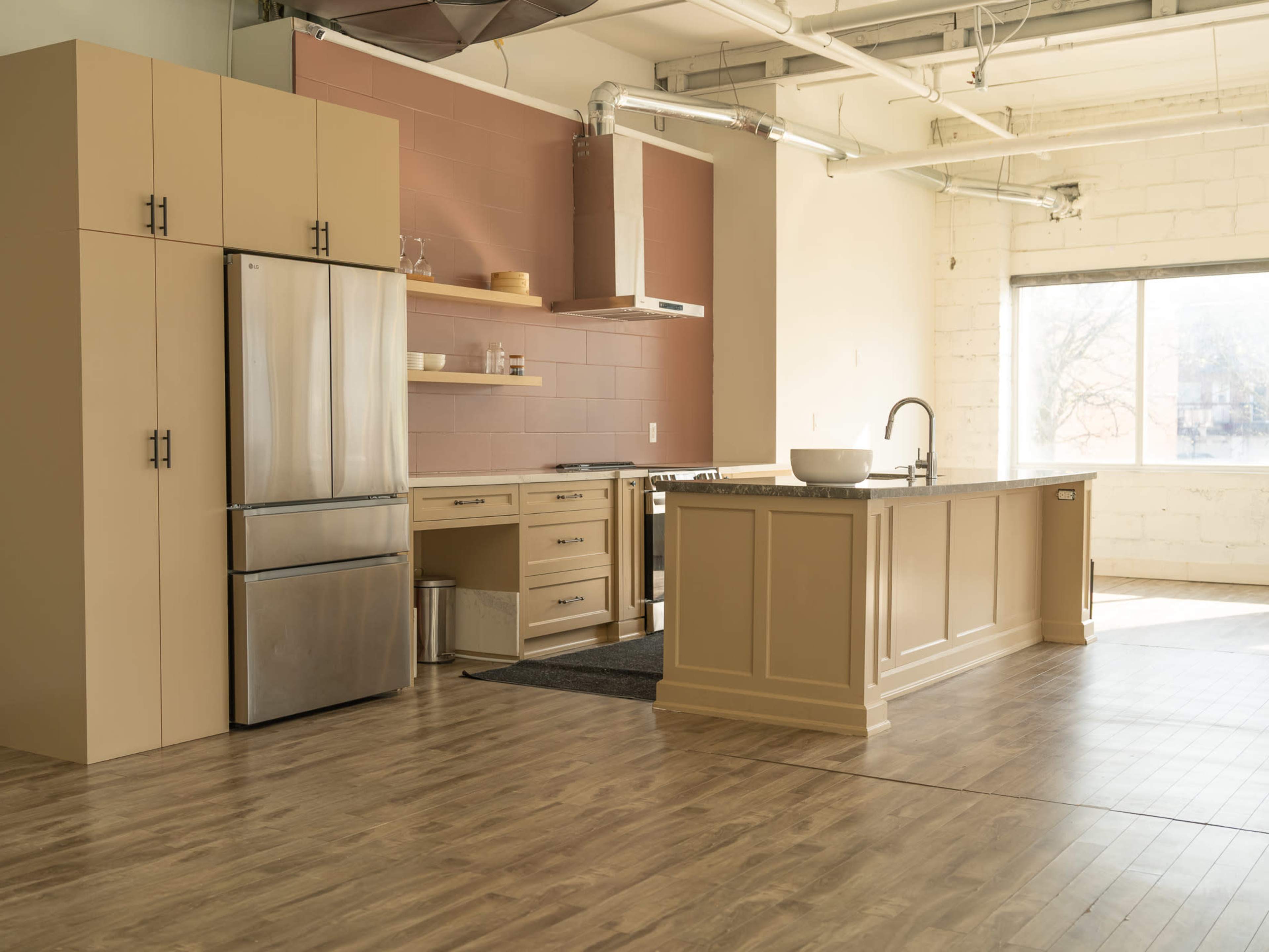 The image shows a modern kitchen with beige cabinetry, stainless steel appliances, and large windows letting in natural light.