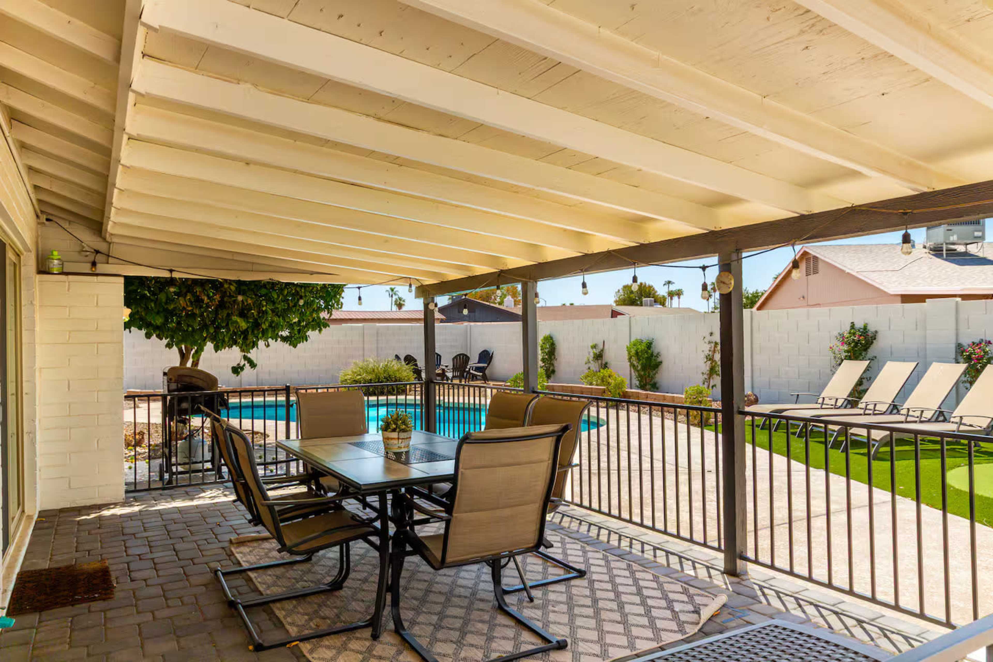 A covered patio with a dining table and chairs overlooks a swimming pool and landscaped yard.