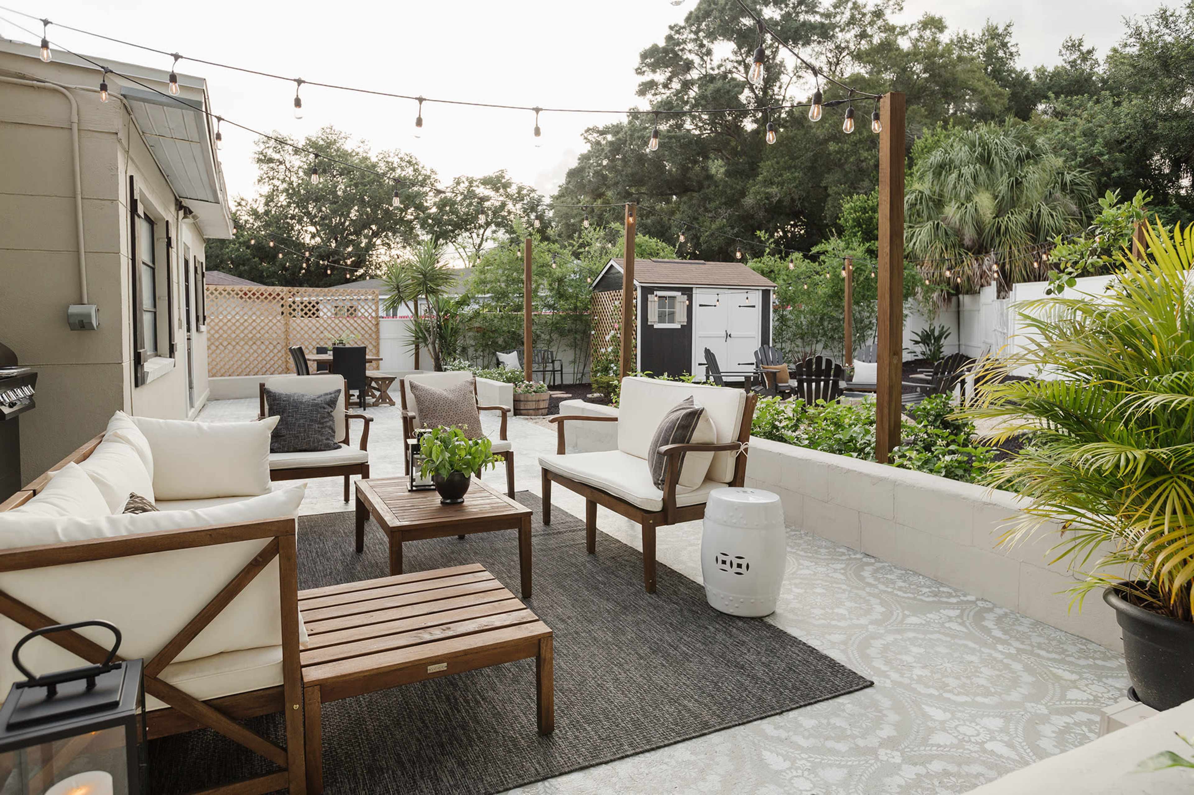 The outdoor area features a seating arrangement with white chairs and wooden tables on a patterned rug, surrounded by greenery and a small shed in the background.