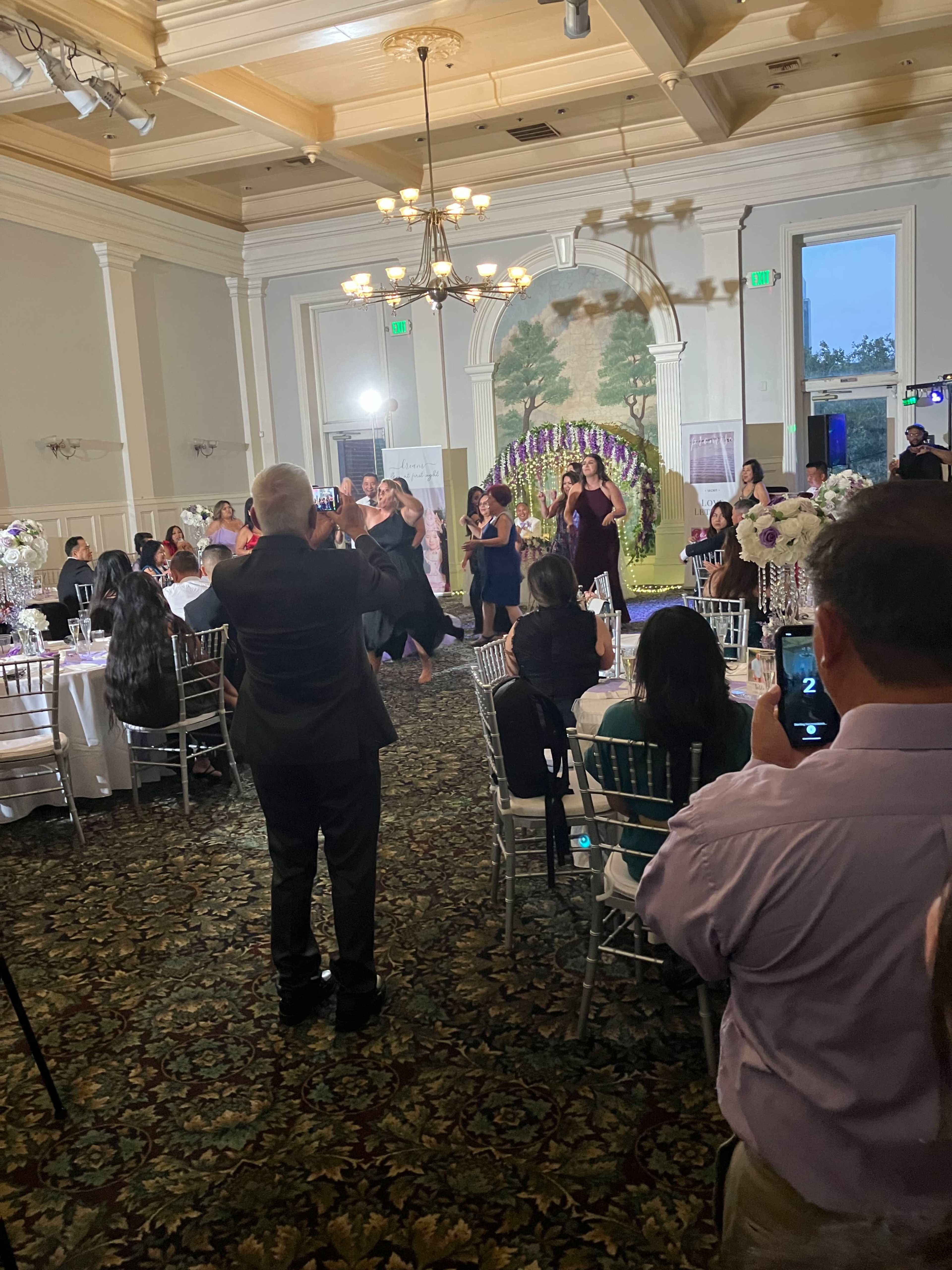 A formal indoor event with guests seated at tables, some taking pictures, as a group of people participate in a dance in the center of the room.