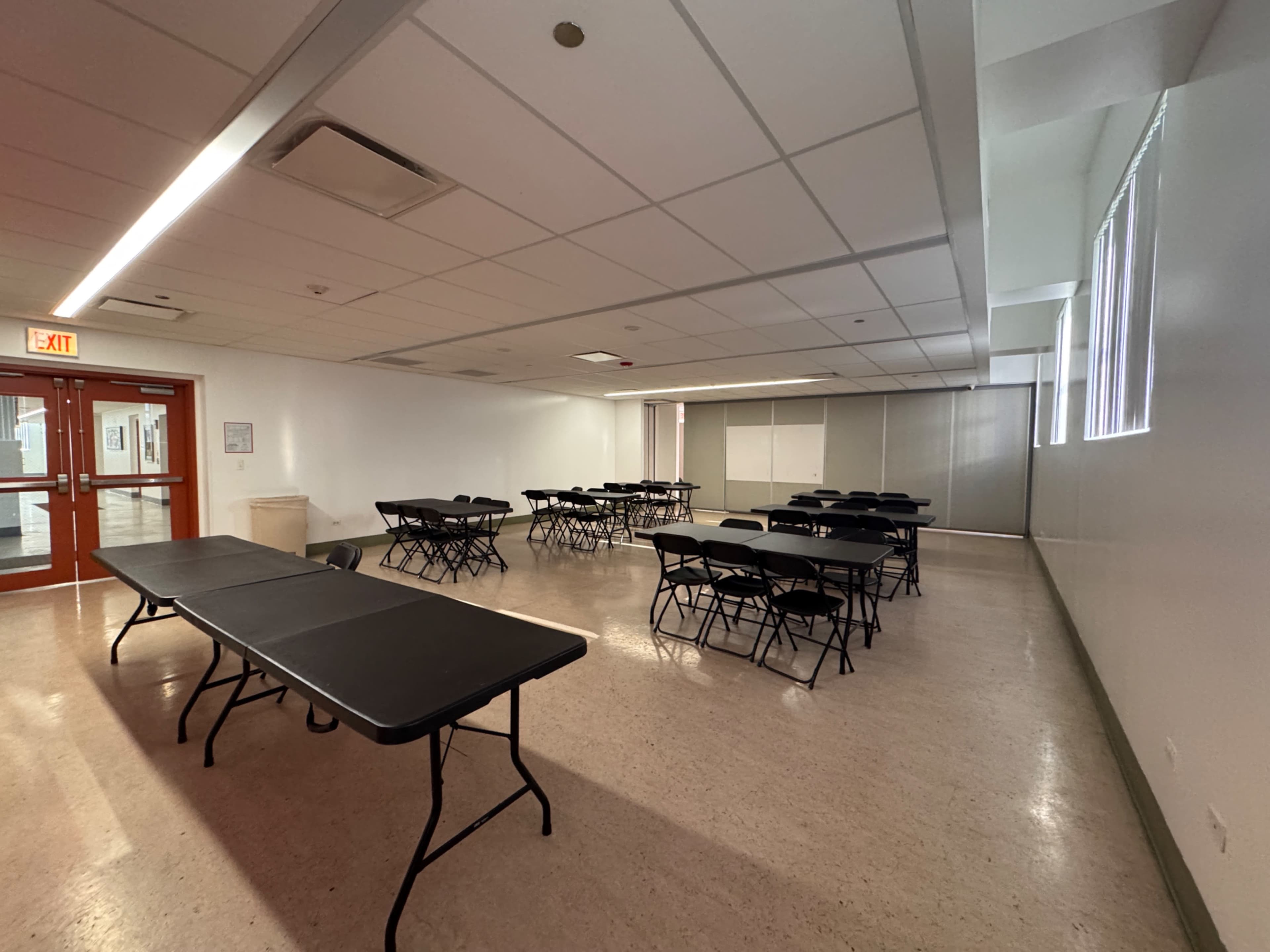 The image shows a spacious room with multiple black folding tables arranged in a grid pattern and a large window along one wall.