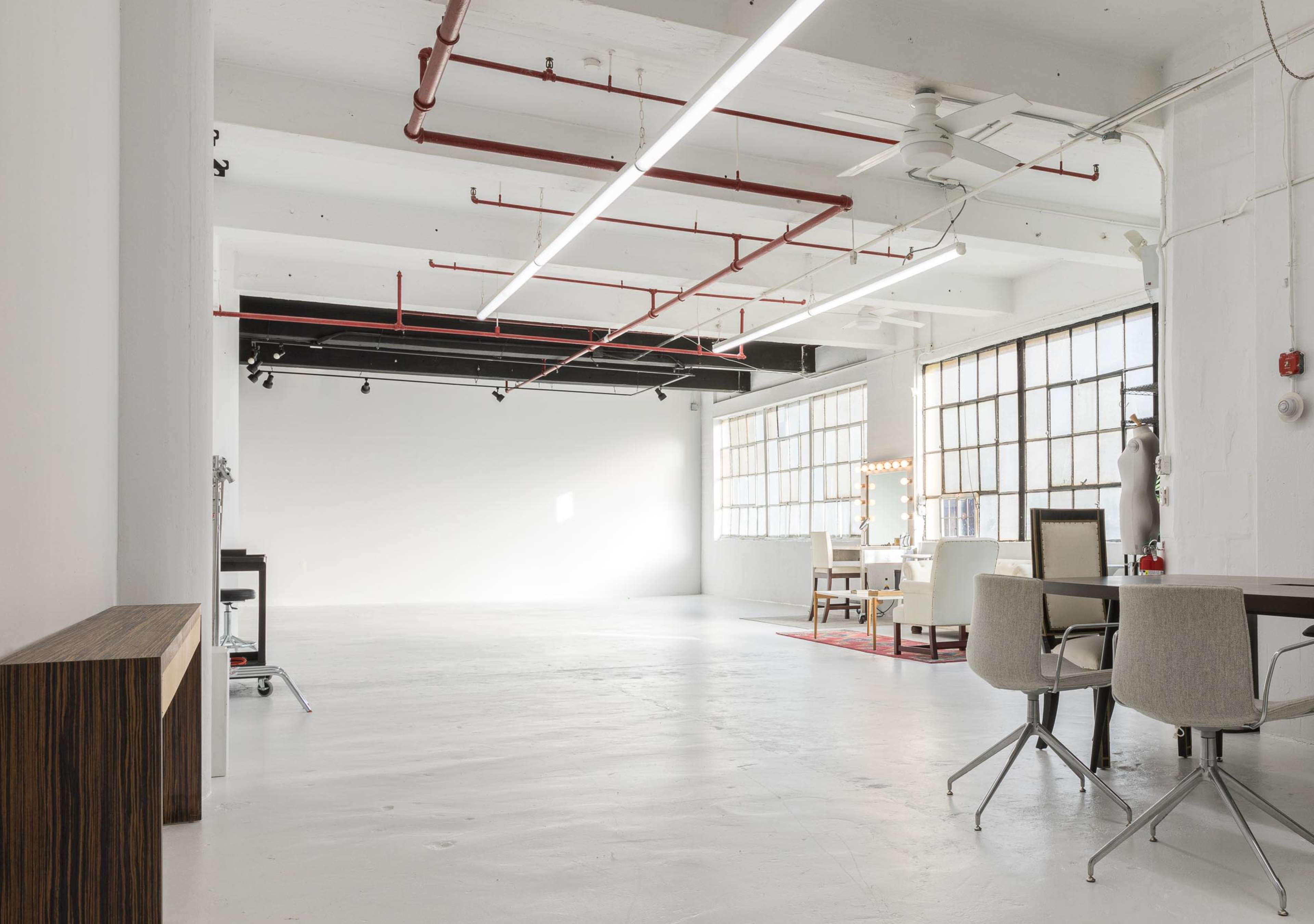 The image shows a spacious, industrial-style room with white walls, large windows, and minimal furniture including a table and chairs.