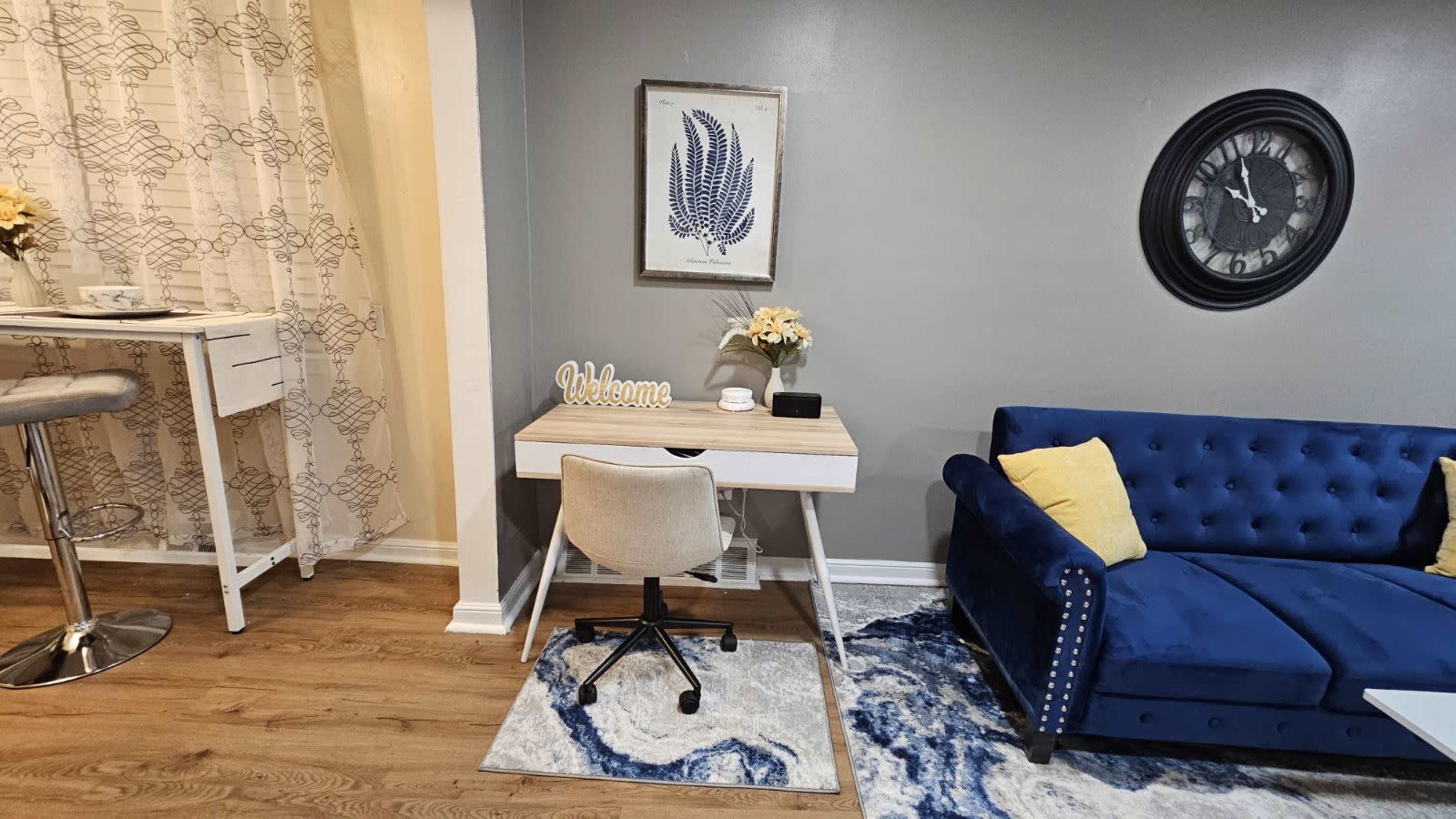A modern living space featuring a blue sofa, a small desk with a chair, a wall clock, and decorative elements like a framed artwork and flowers.