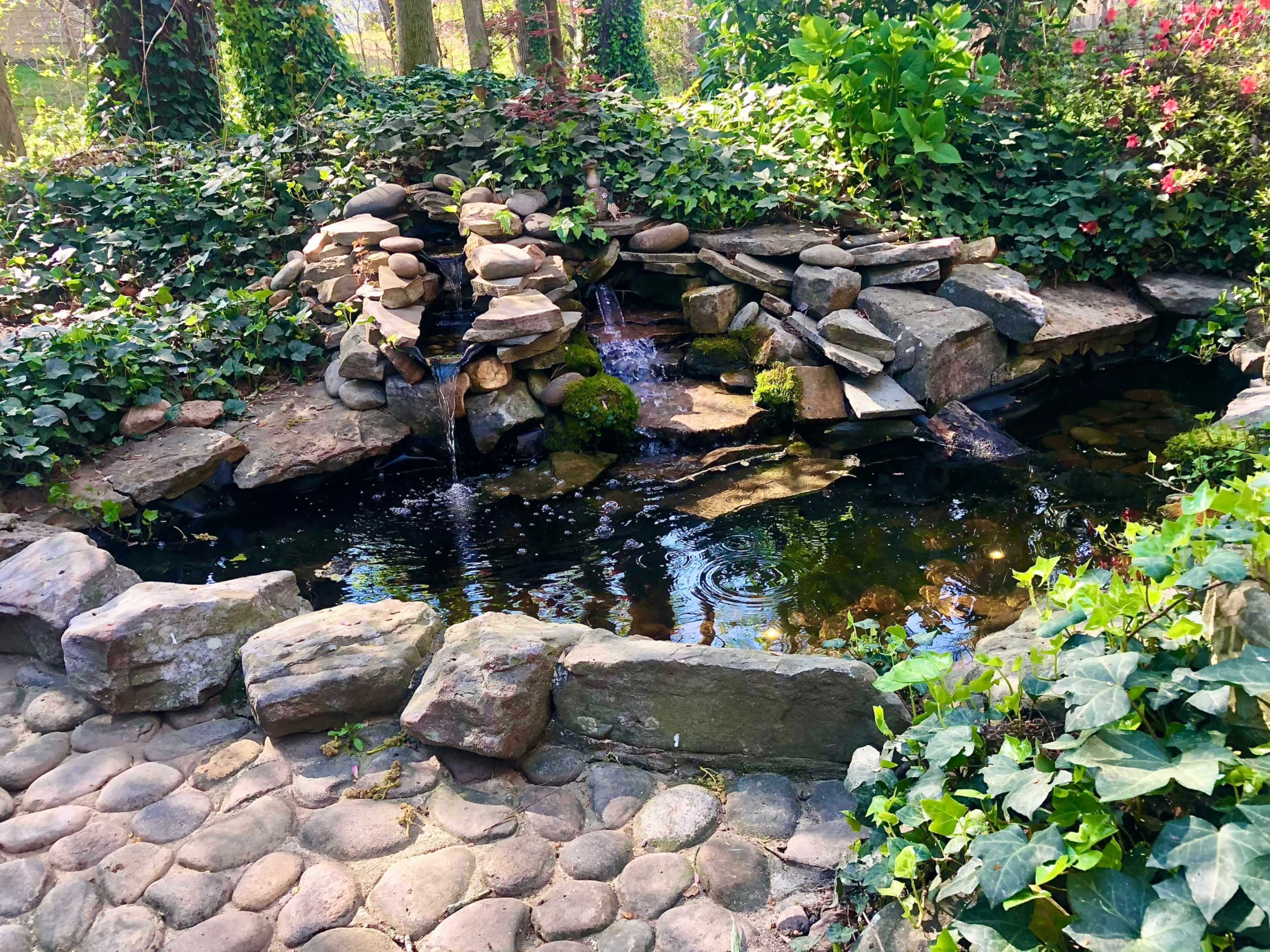 A natural stone fountain surrounded by greenery and a pond with rippling water.