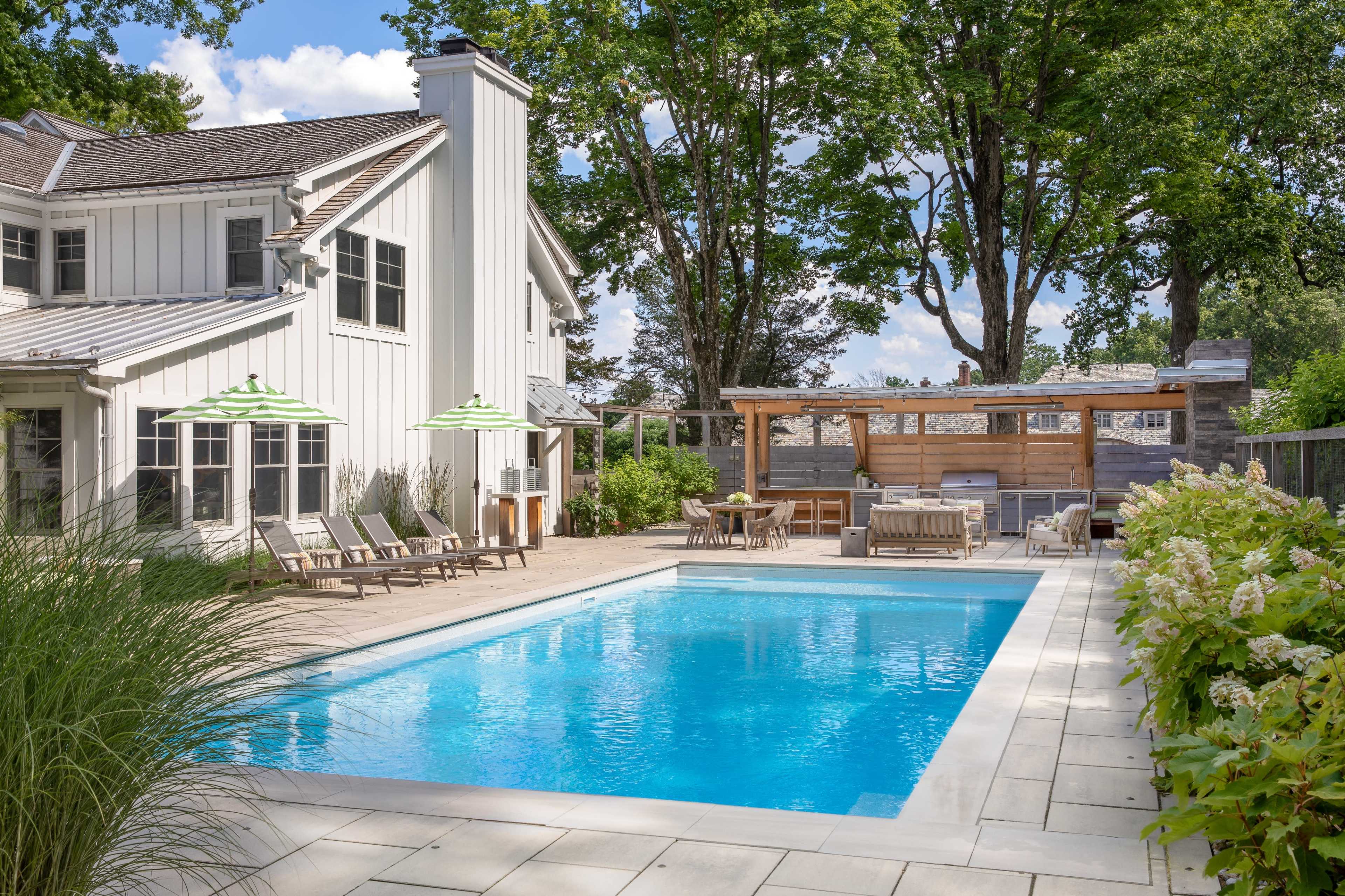 A backyard features a rectangular swimming pool surrounded by lounge chairs, a covered seating area, and lush greenery.