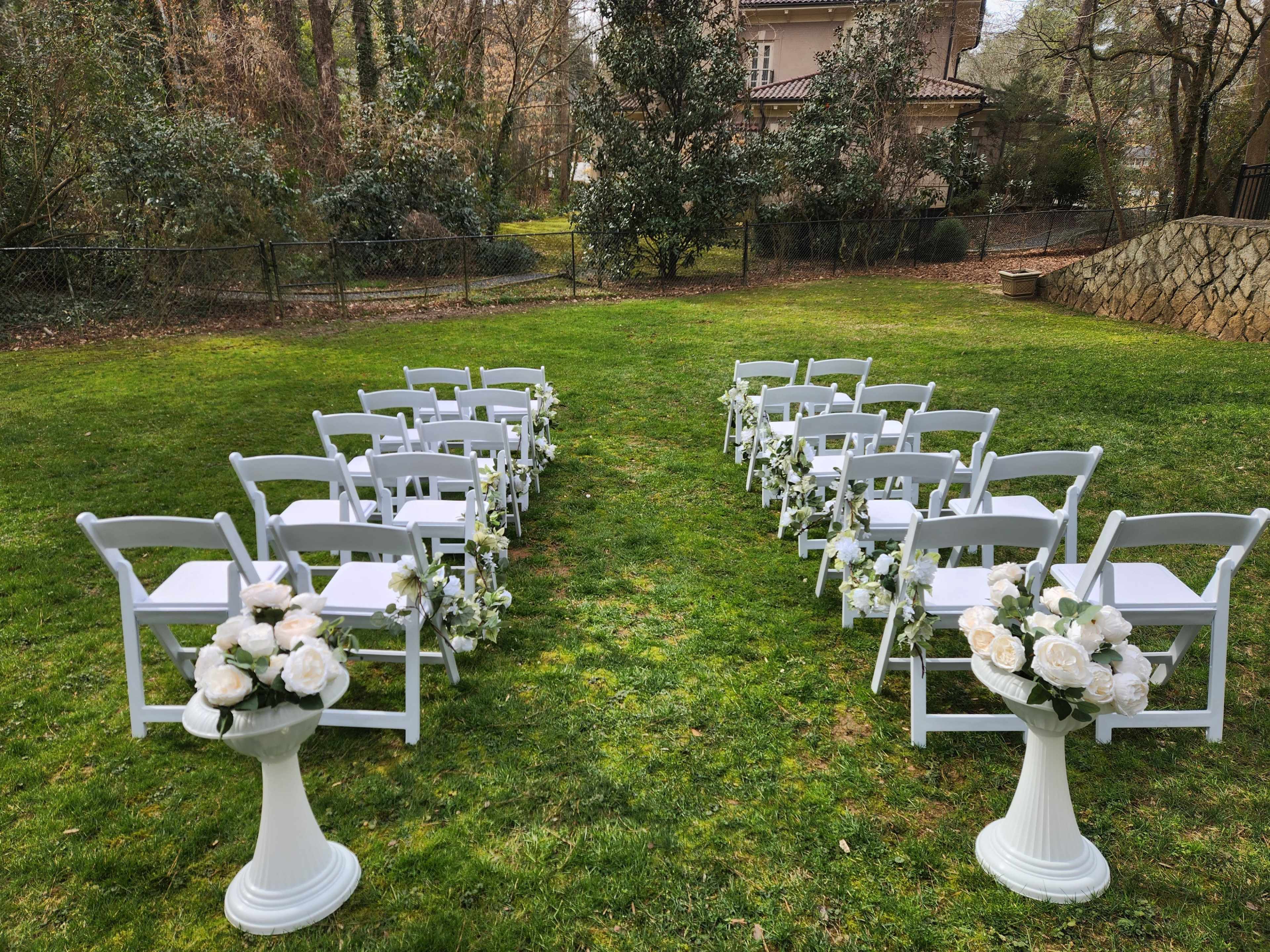 The scene shows rows of white chairs arranged in a grassy area, with floral arrangements on pedestals at each end, set for a wedding ceremony.