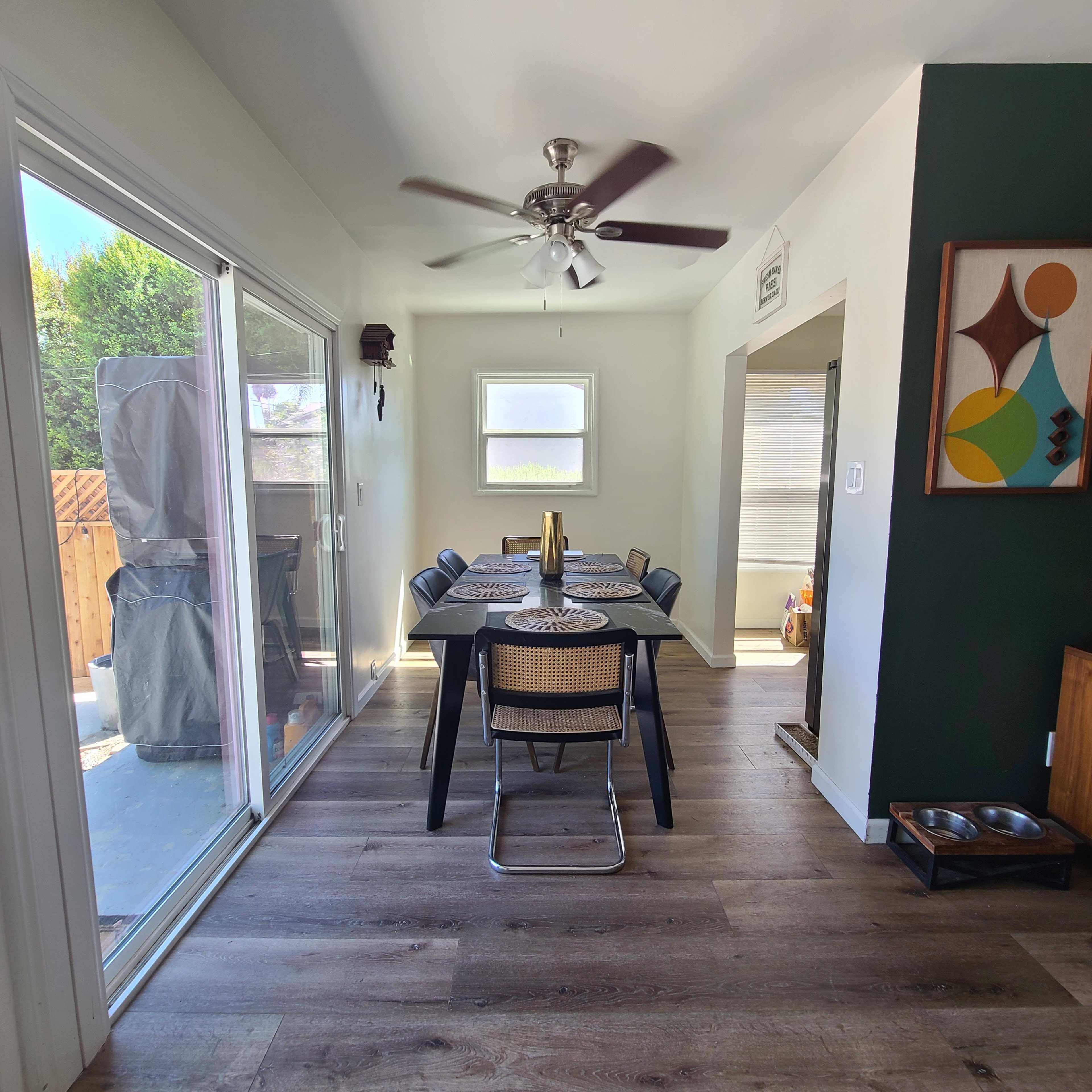 The image shows a modern dining area with a black table set for six, featuring a ceiling fan and large windows that let in natural light.