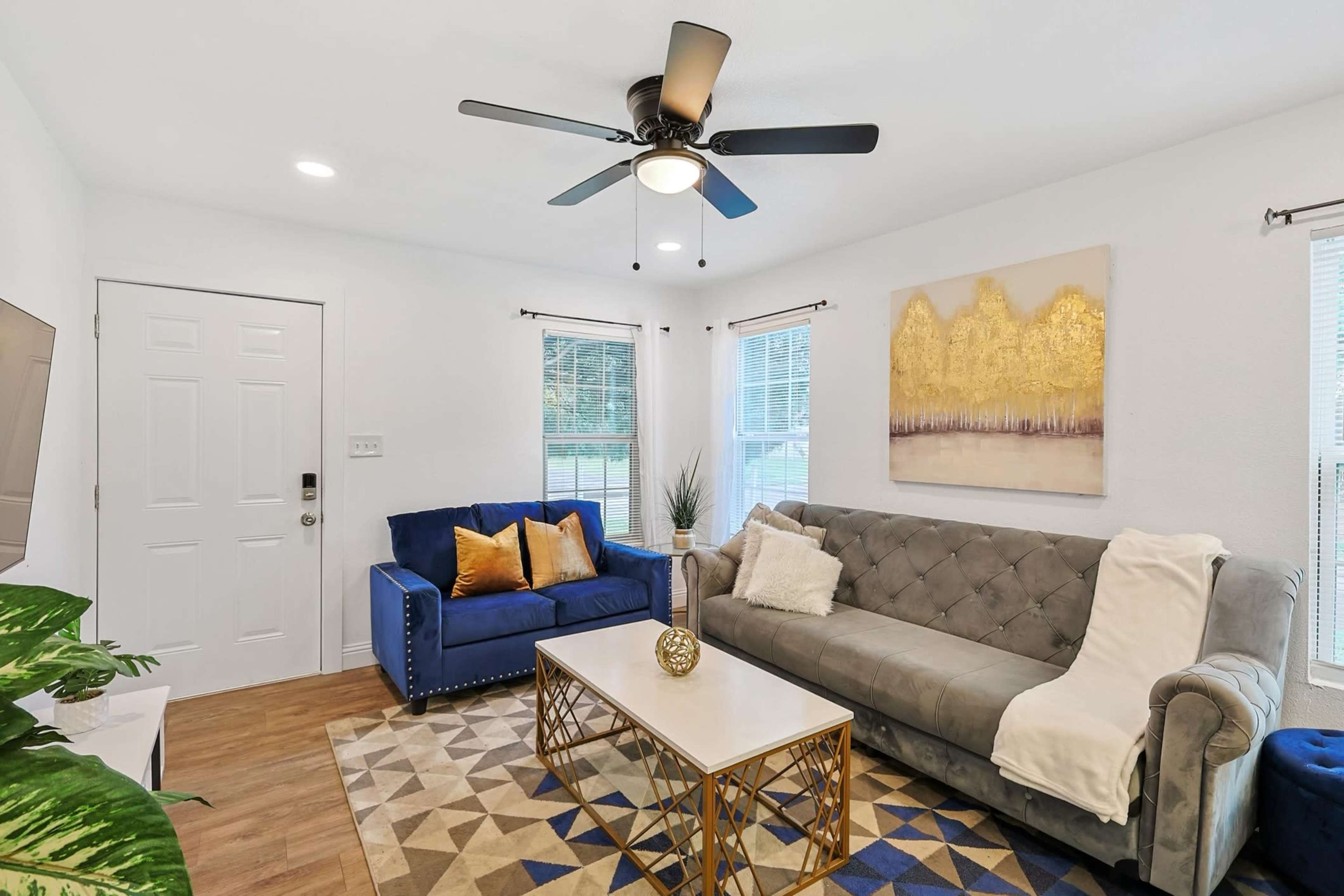 The living room features a blue sofa and a gray couch facing a coffee table on a geometric patterned rug, with a ceiling fan and light fixtures above.