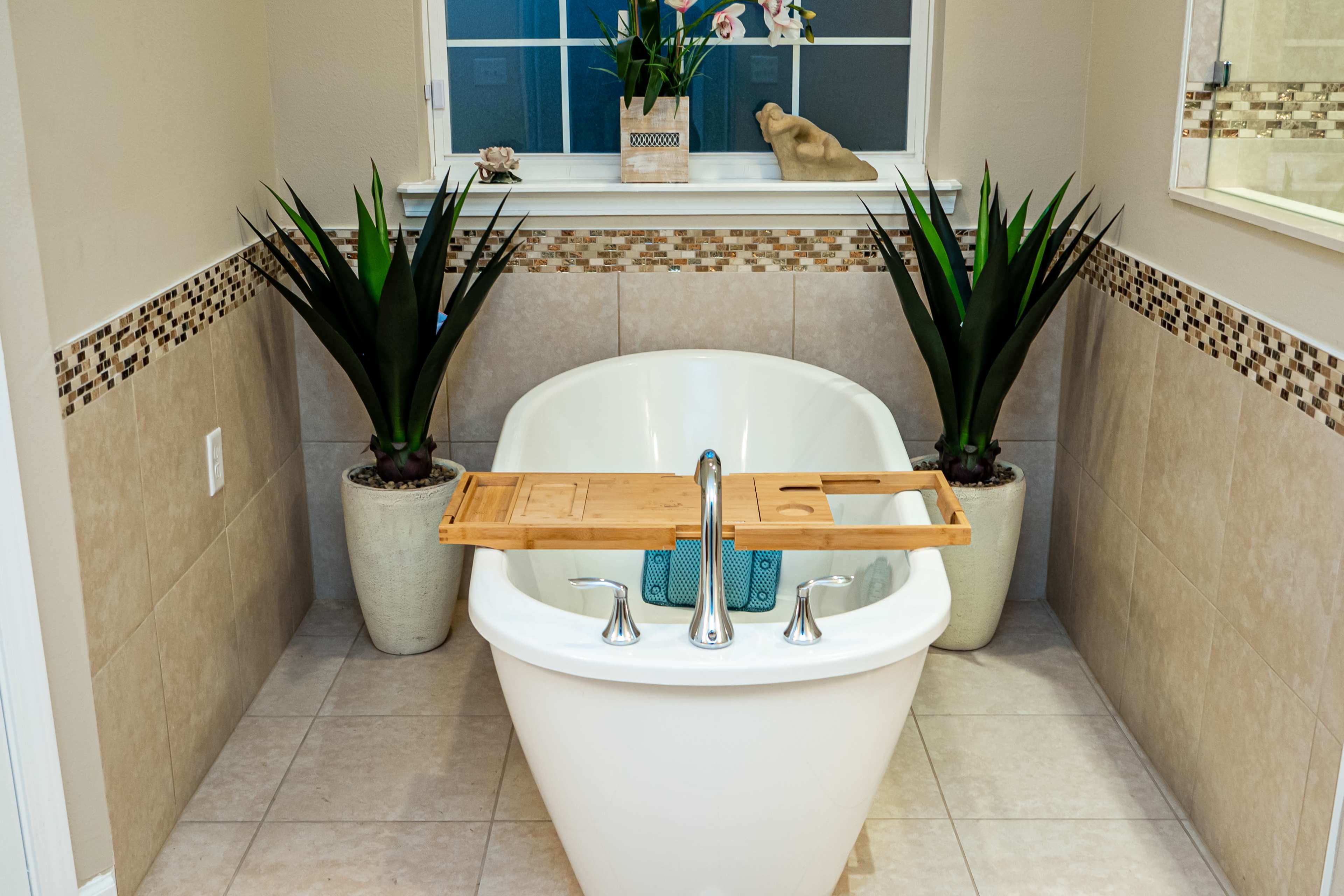 A freestanding white bathtub is centered in a neatly tiled bathroom, flanked by two tall plants and featuring a wooden tray across the top.