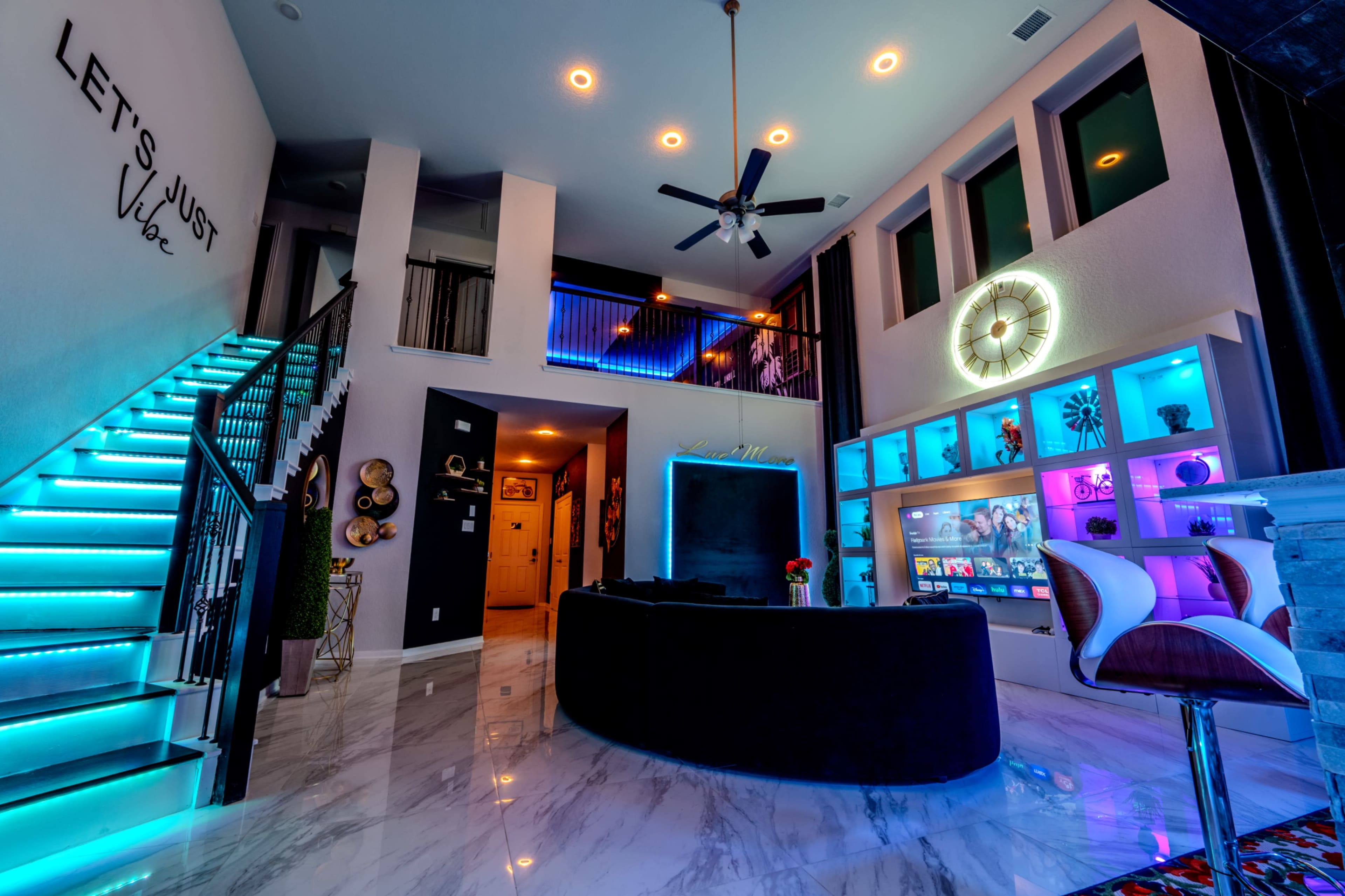 The image shows a modern living space featuring a sleek black couch, a glowing entertainment unit, a staircase with decorative lighting, and vibrant wall accents.