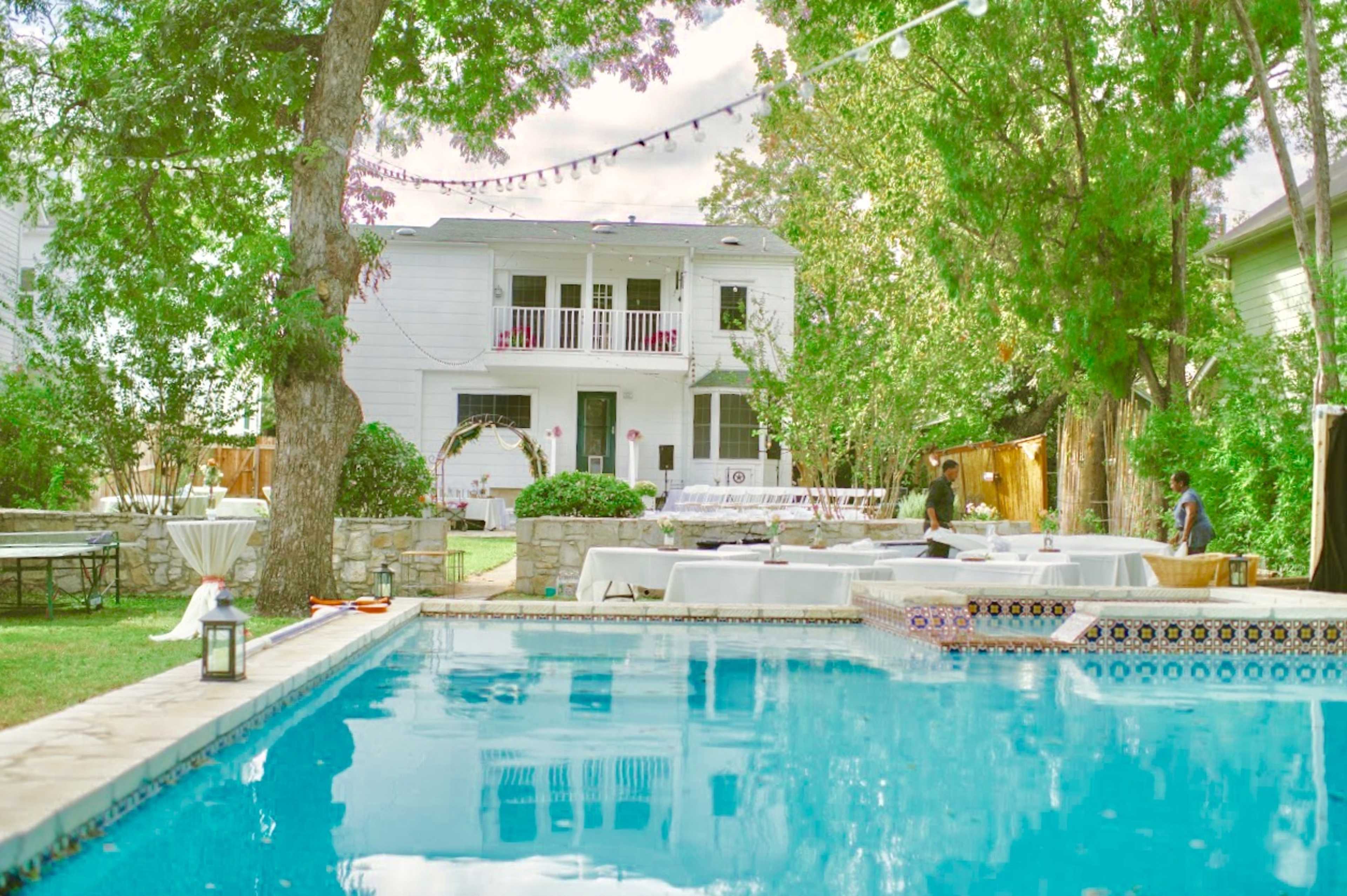 A clear swimming pool reflects a two-story house surrounded by greenery and outdoor seating areas for gatherings.
