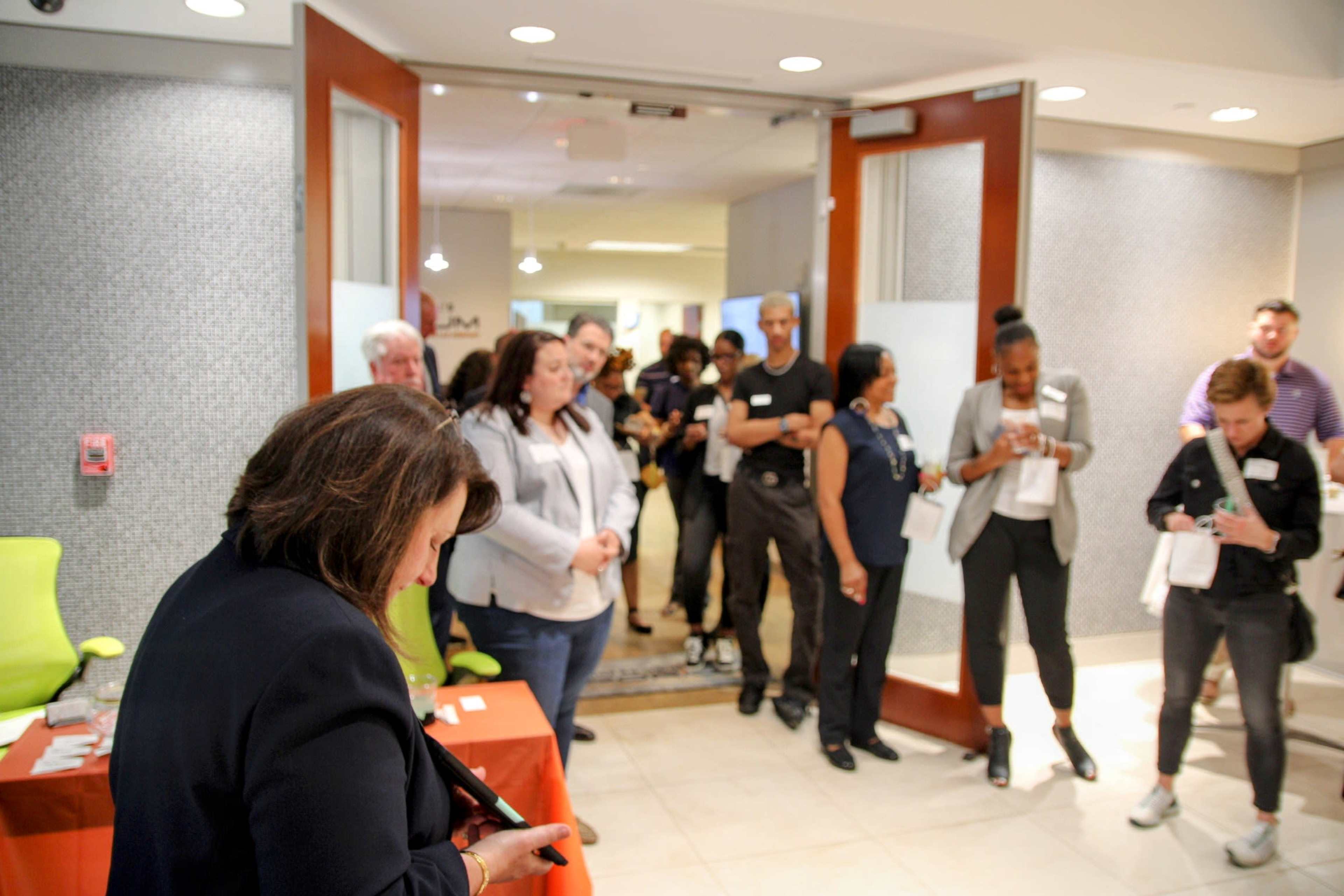 A group of people stands in line inside a building, with some individuals interacting with their smartphones and others engaged in conversation.