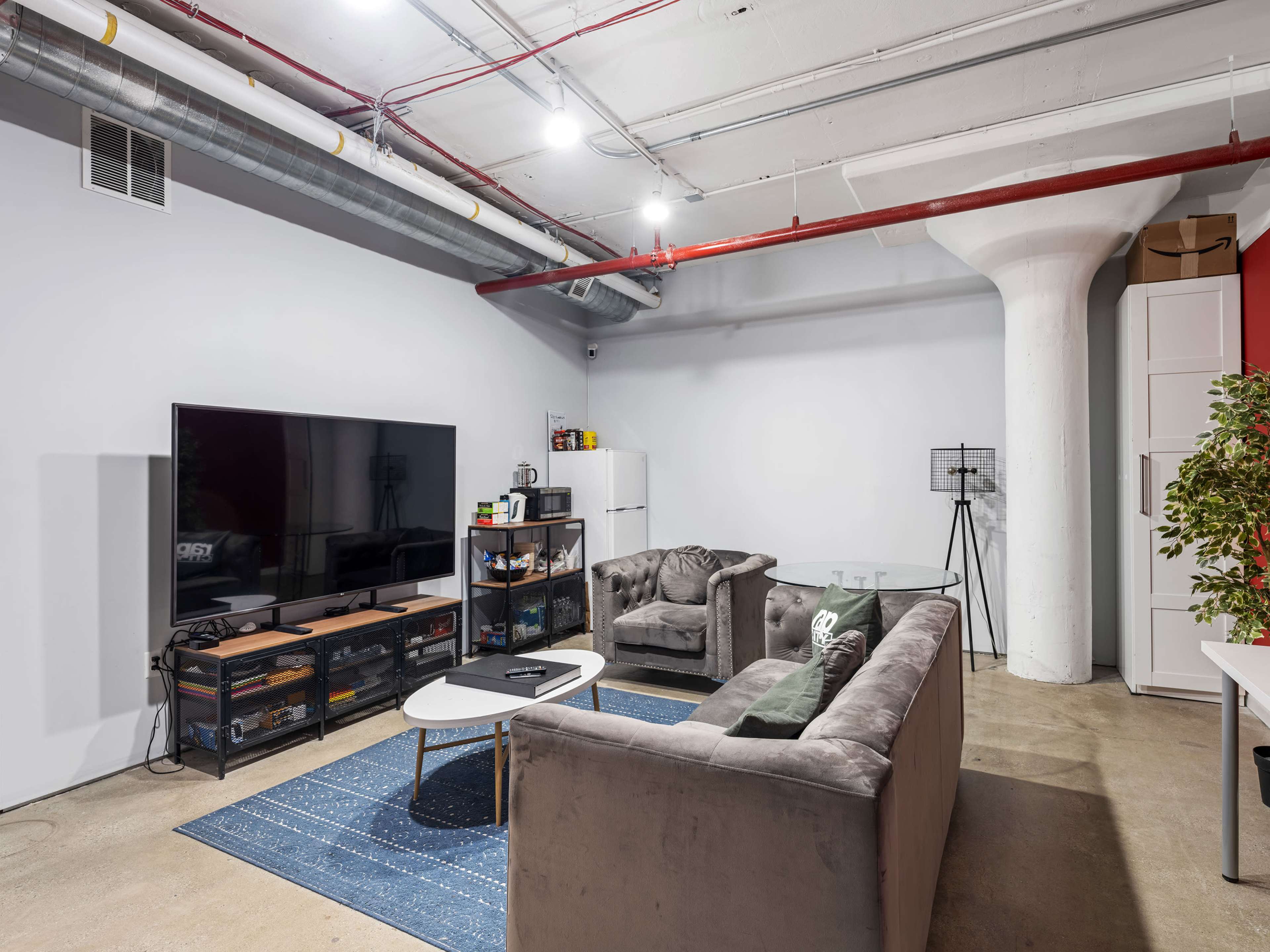 A spacious living room features a large television, two gray sofas, a coffee table, and a white refrigerator against one wall.
