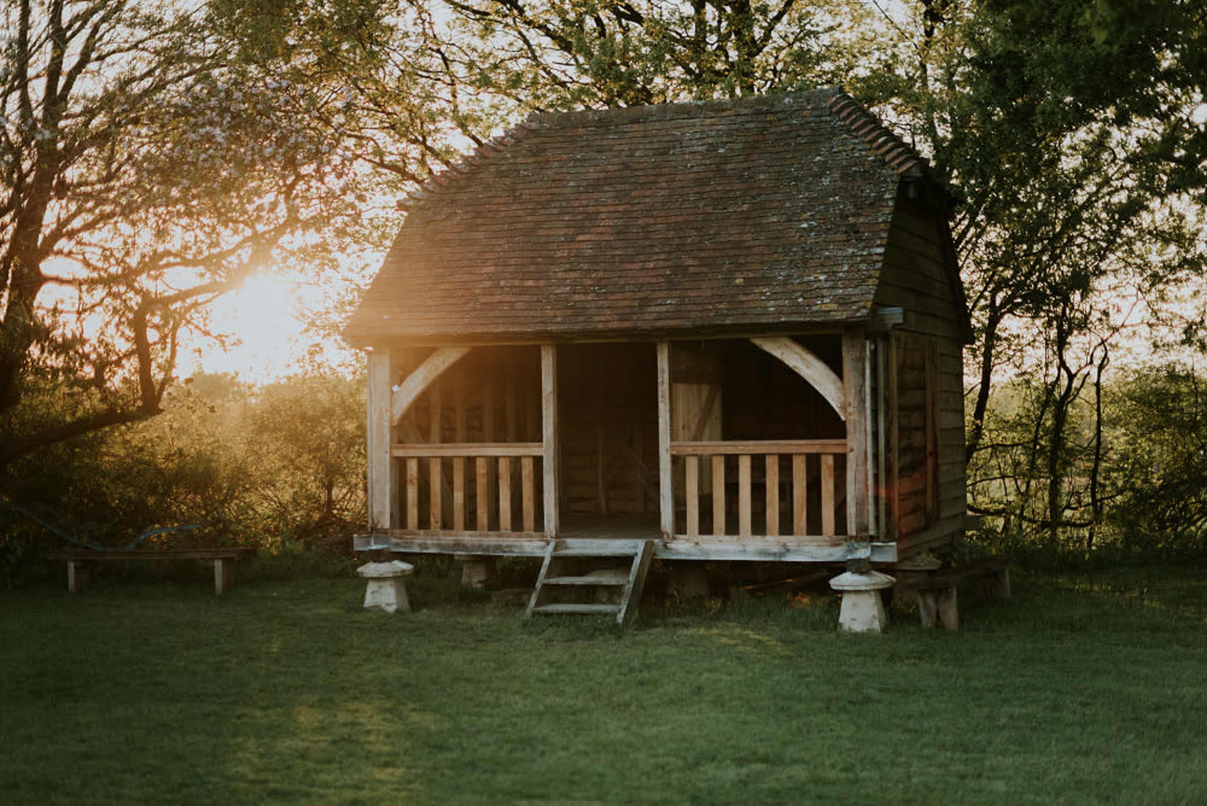 A wooden cabin with a slanted roof and a front porch stands in a grassy area surrounded by trees at sunset.