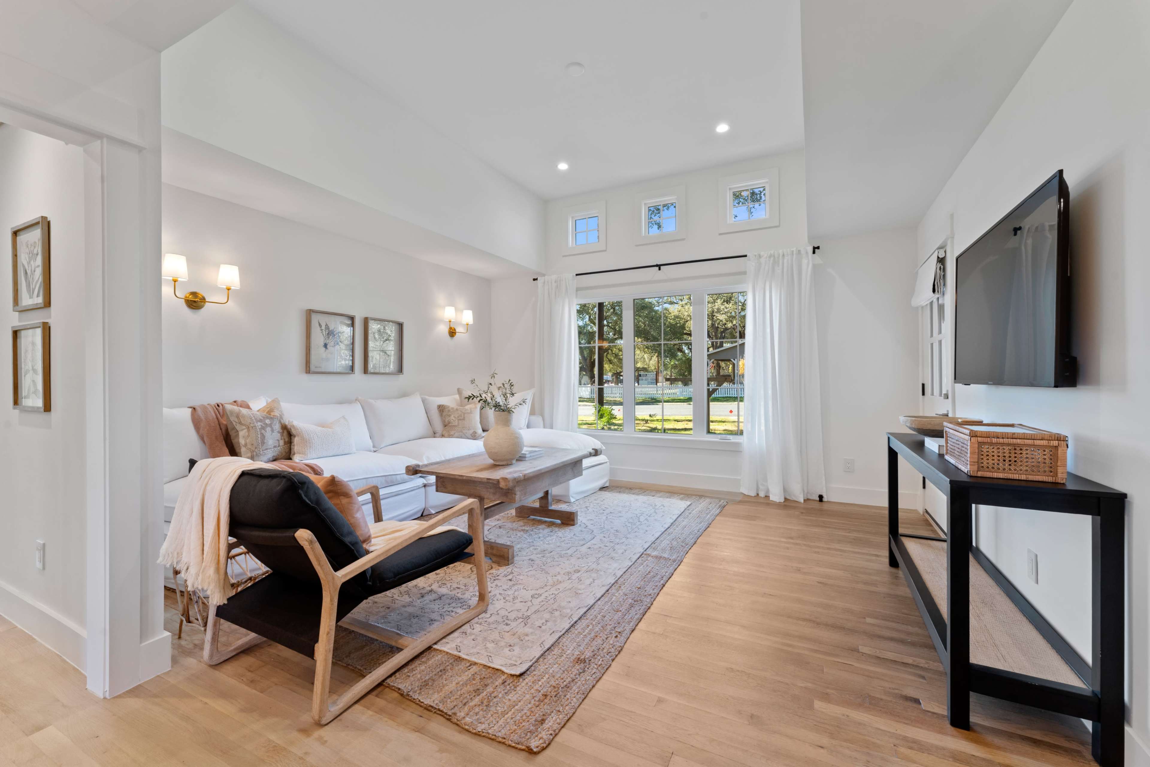 A bright living room features a white sofa, a wooden coffee table, a black chair, and large windows overlooking a garden.