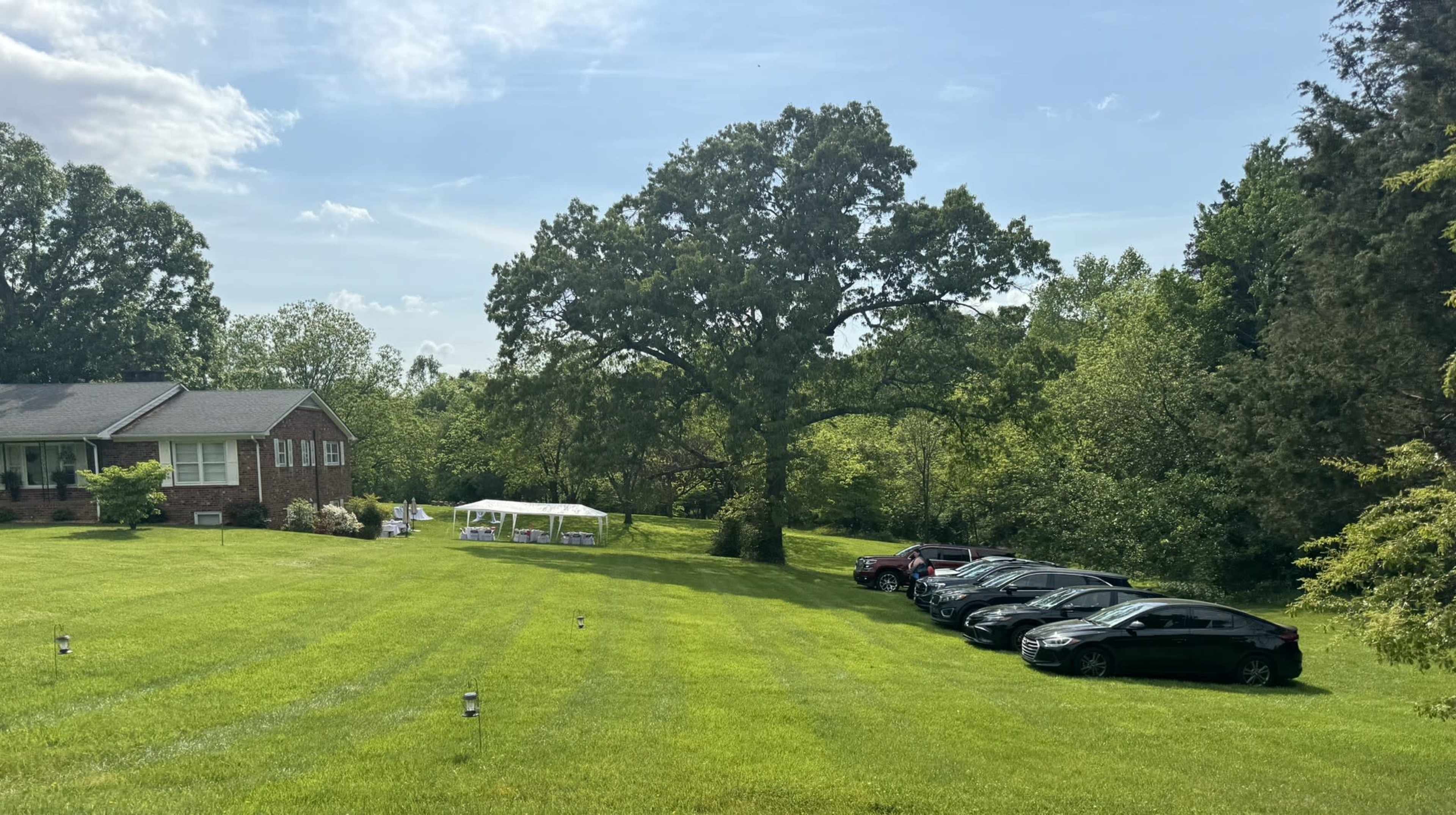 A large brick house sits on a well-maintained lawn with several parked cars lined up beside a white tent in the background.