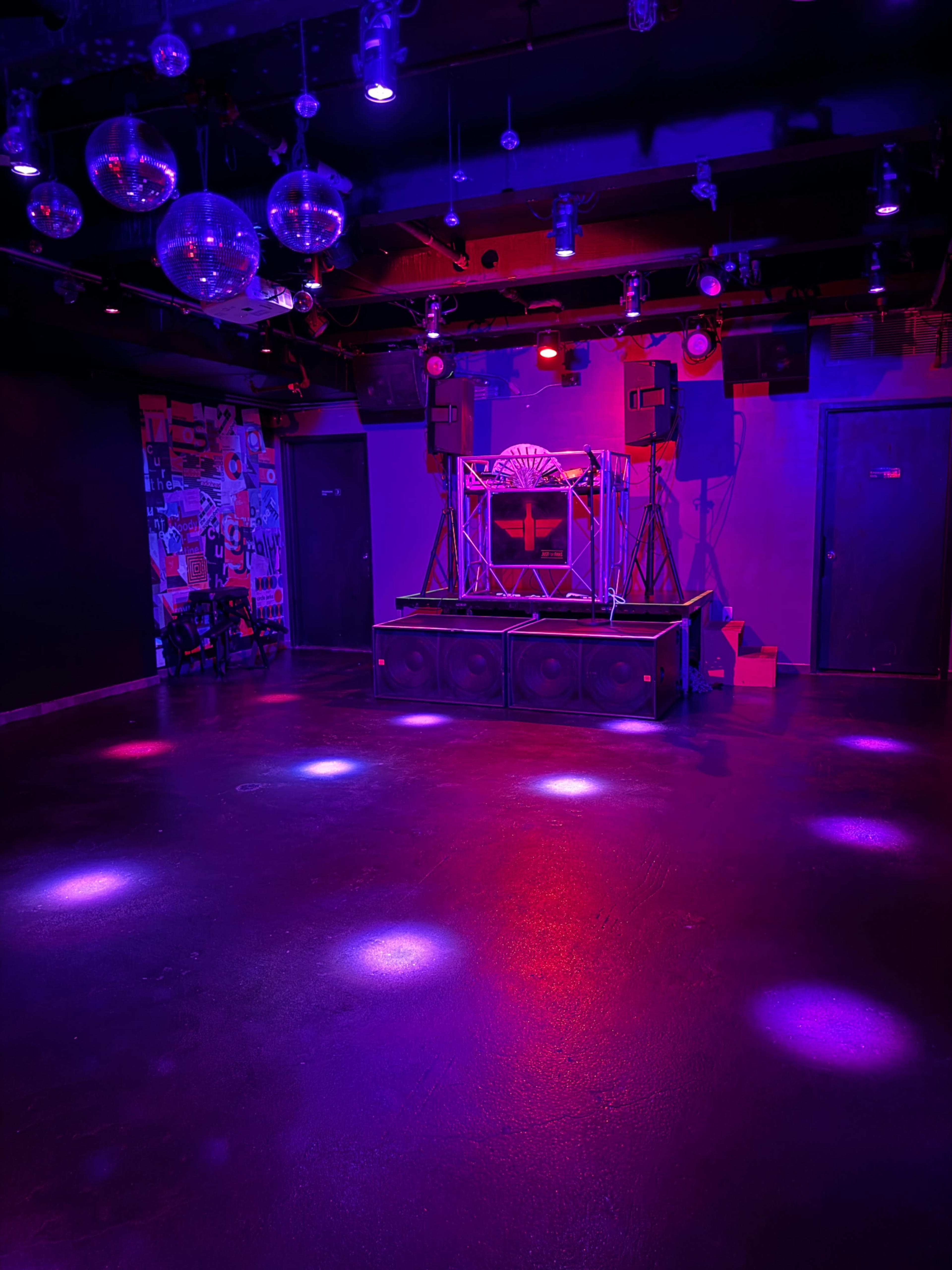 A dimly lit music venue with a raised stage, sound equipment, and disco balls hanging from the ceiling.