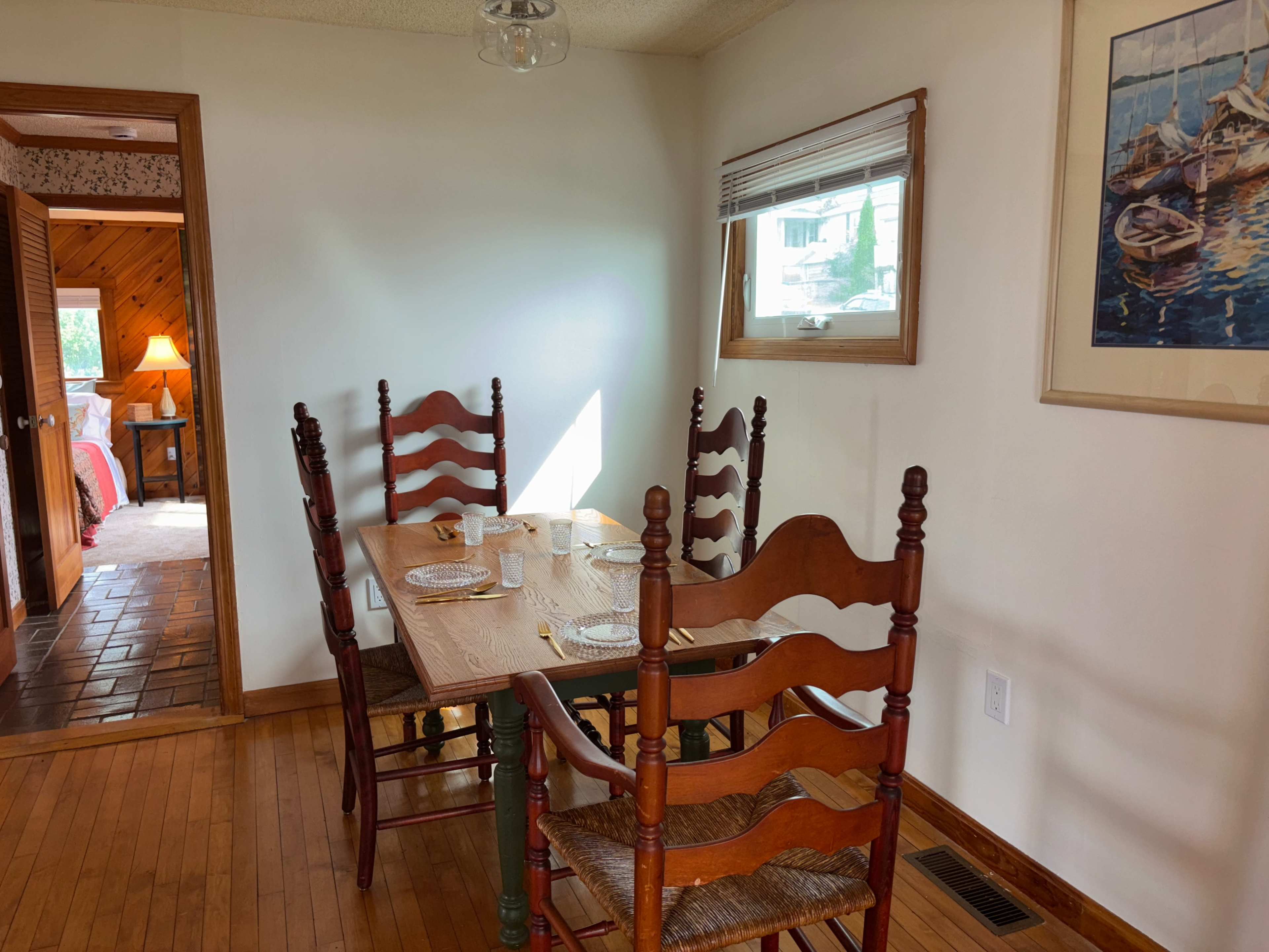 A wooden dining table is set for four, surrounded by tall chairs, in a brightly lit room with a painting on the wall and a doorway leading to another room.