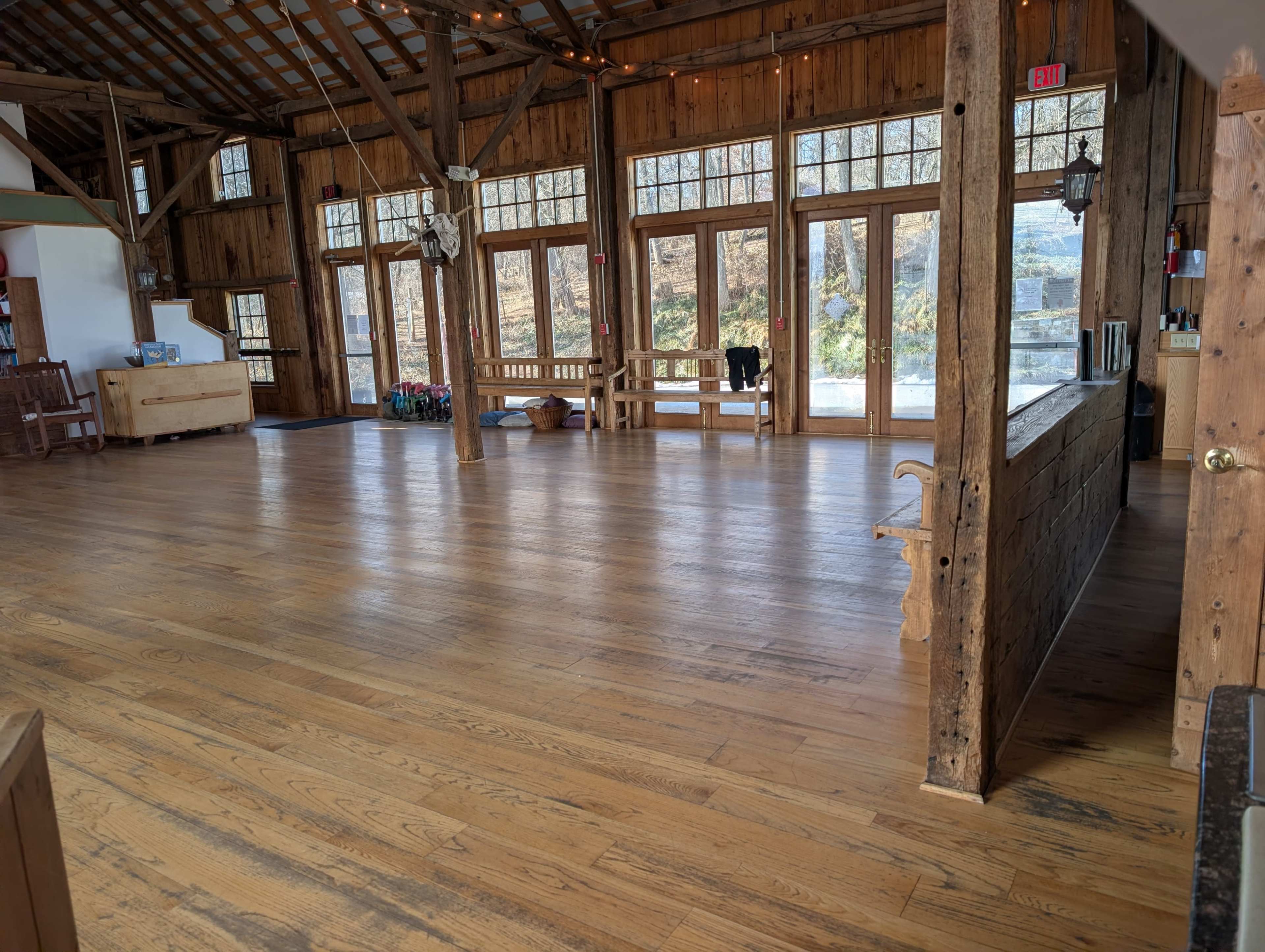 The image shows a spacious, wooden interior of a rustic venue with large windows and exposed beams, featuring a polished wooden floor and a few benches.