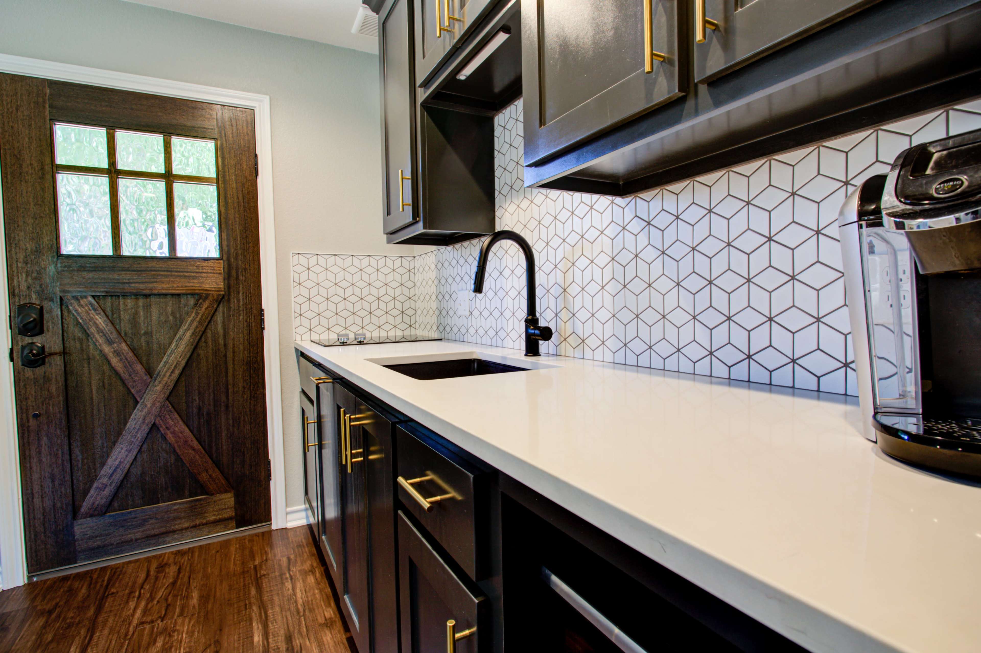 A kitchen features dark cabinets, a white countertop, a geometric backsplash, and a coffee maker next to a wooden door.