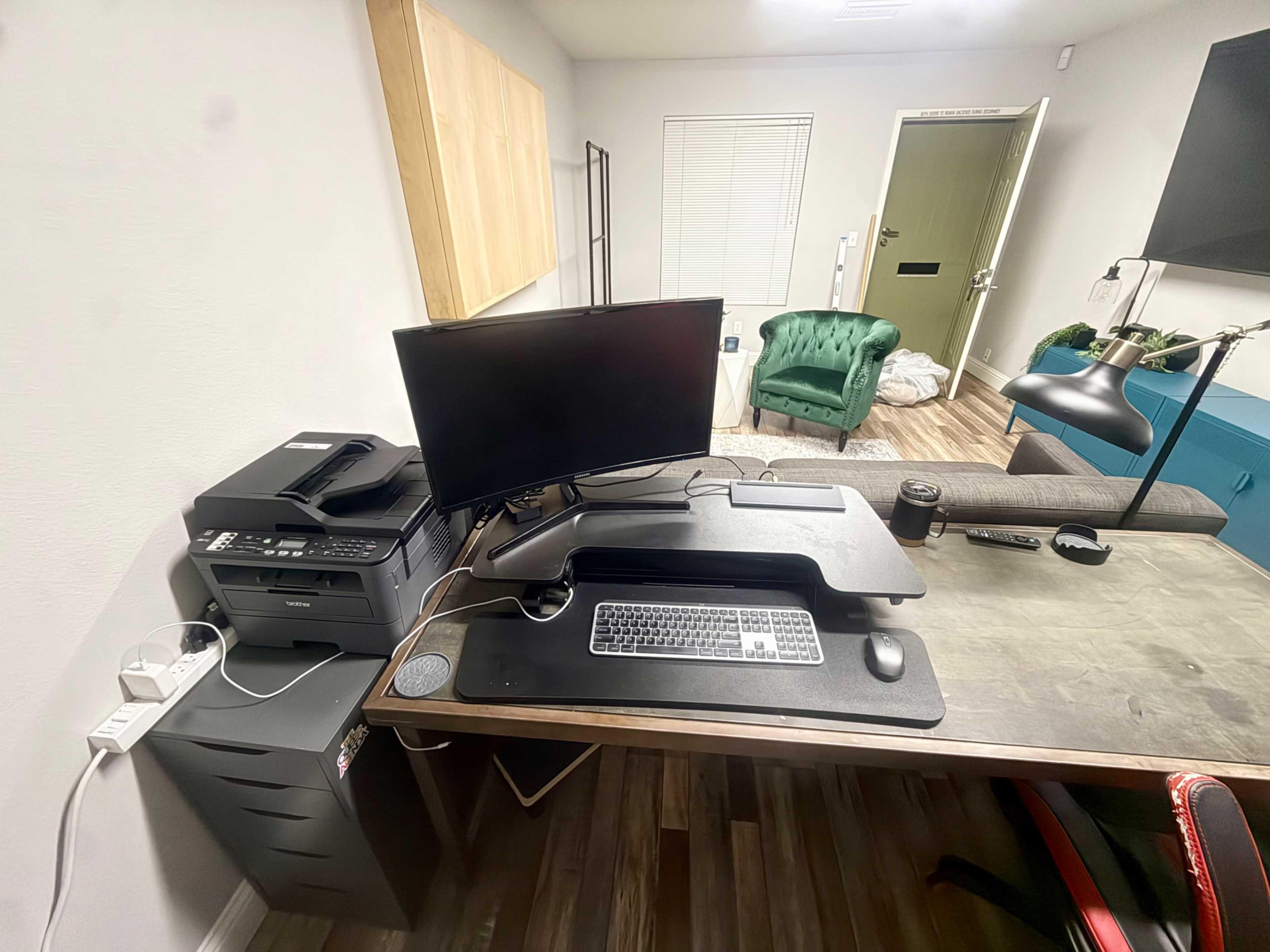 A workspace features a computer monitor, keyboard, and printer on a desk, with a green chair and door visible in the background.