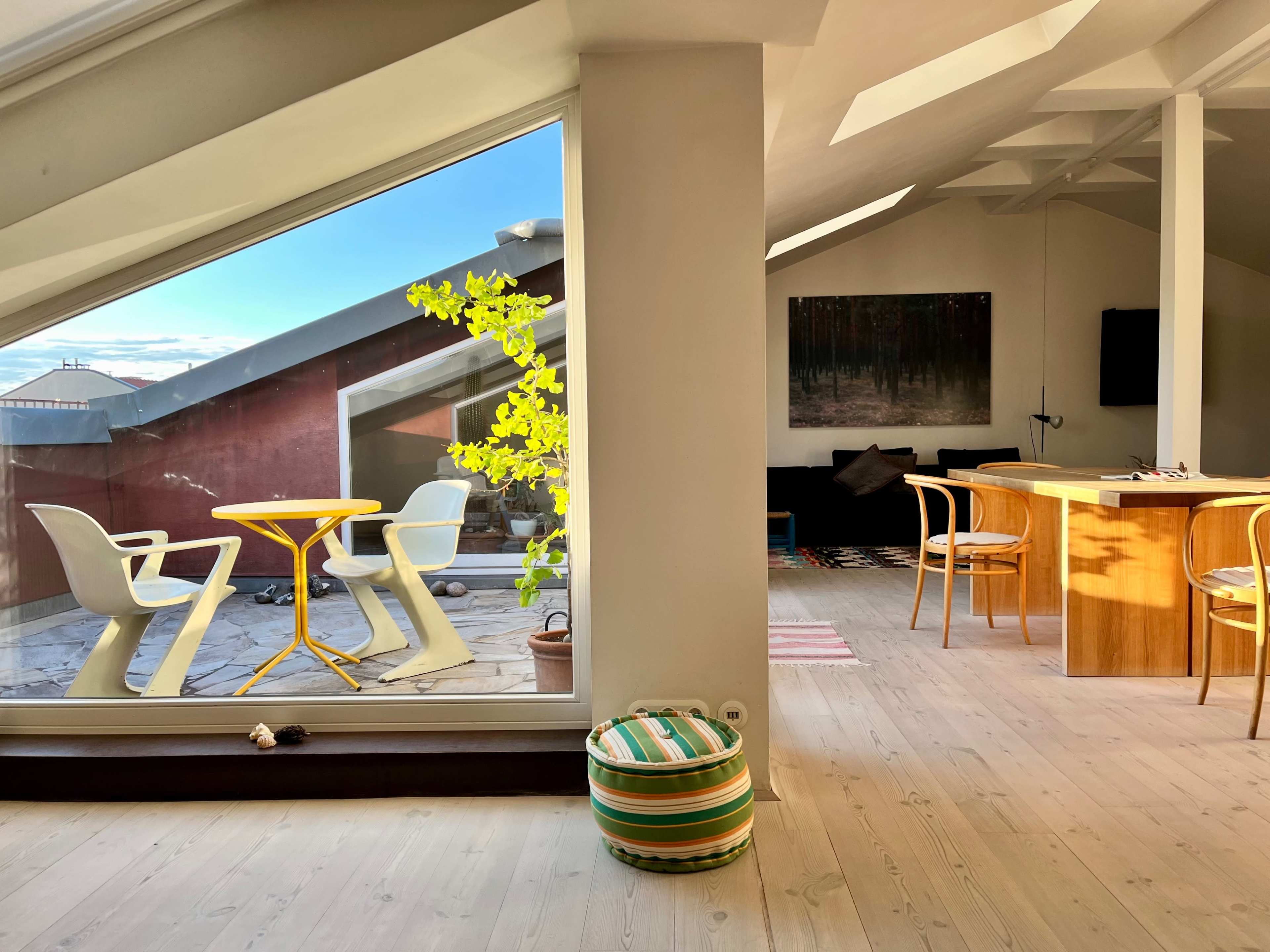 A modern interior with a sunlit dining area featuring a wooden table, and a view of a rooftop terrace with a yellow table and chairs visible through a large window.