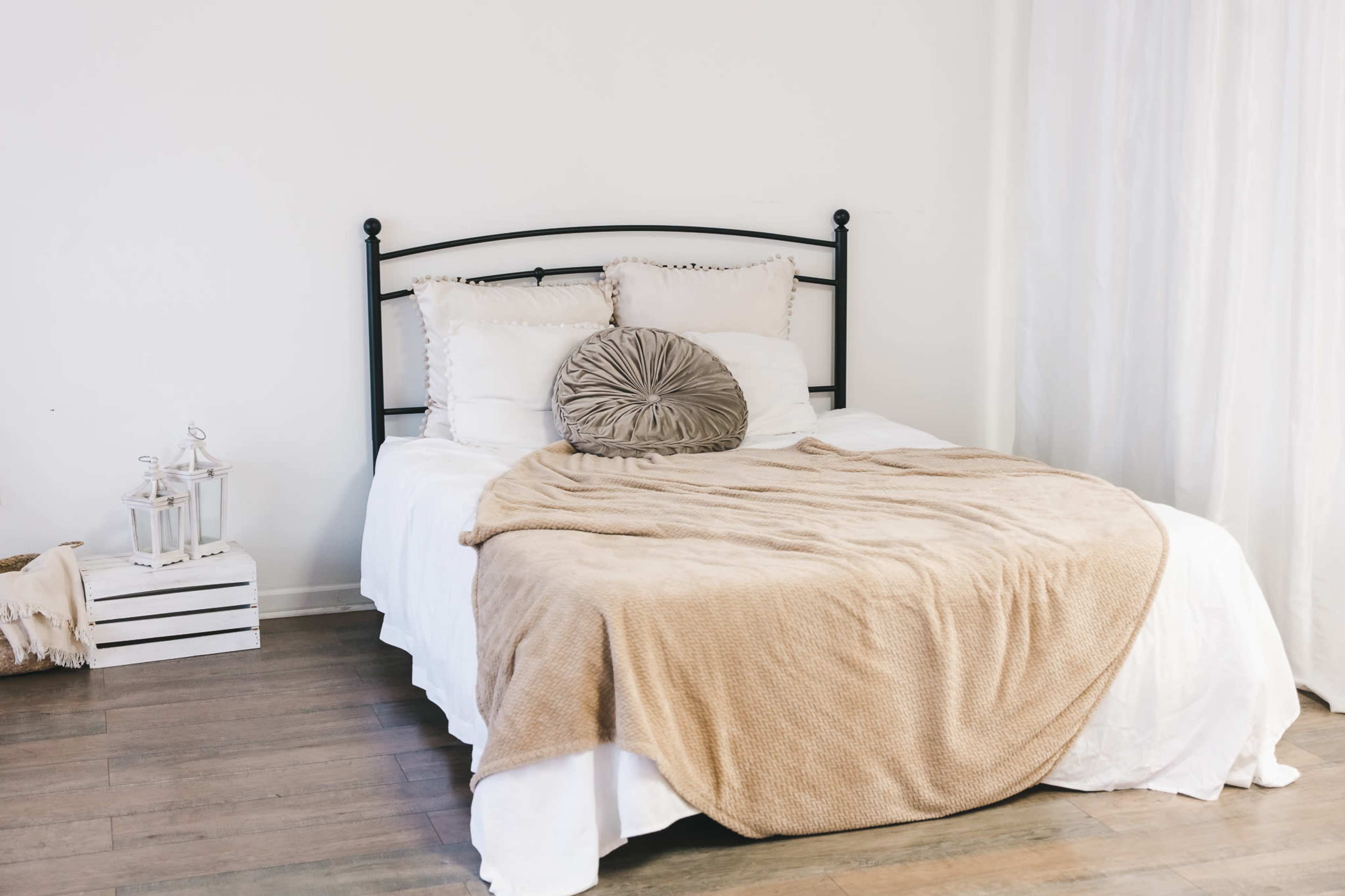 The image shows a neatly made bed with white bedding and a beige throw blanket, accompanied by decorative pillows and minimal decor in a bright, simple room.