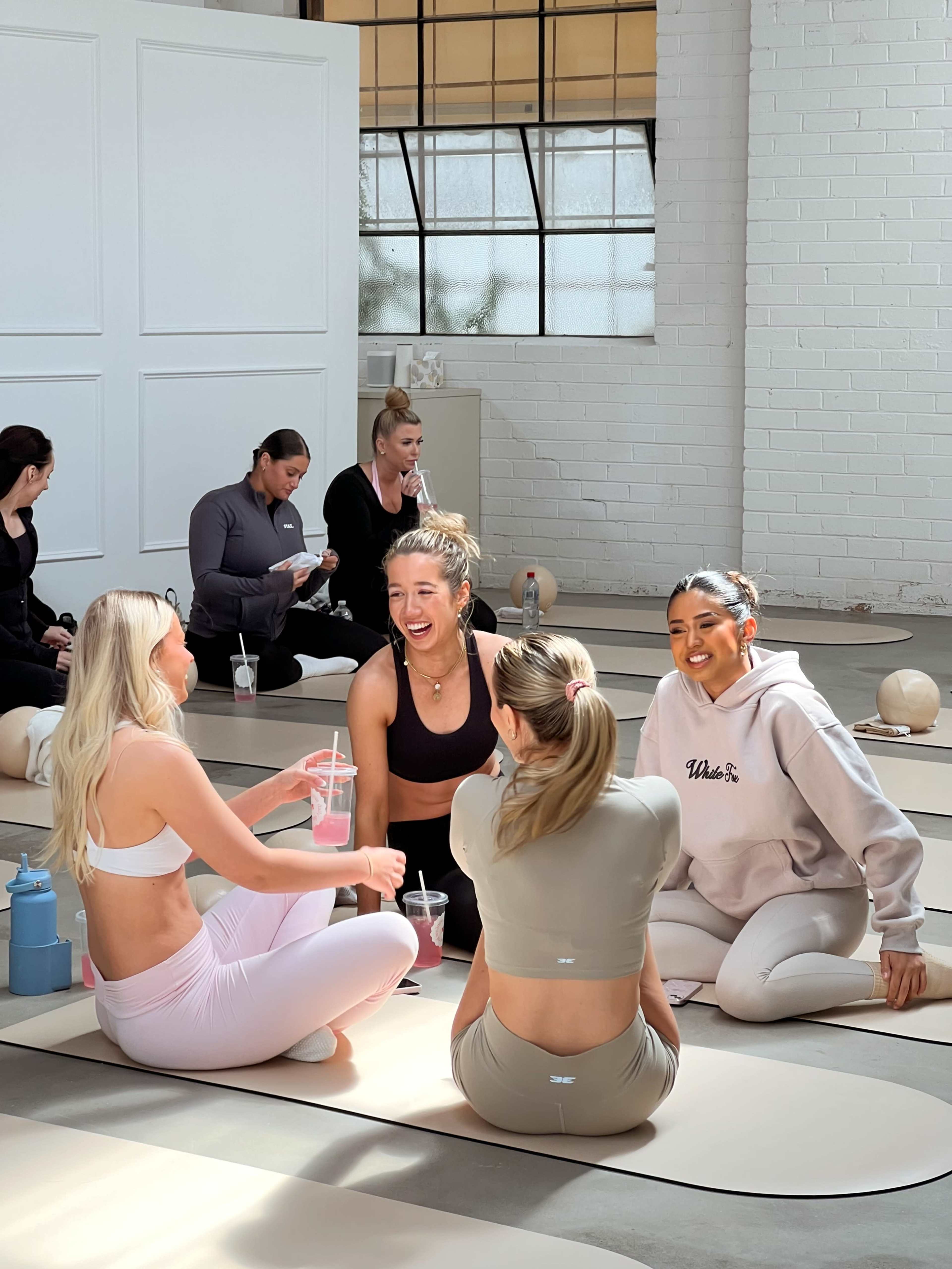 A group of women are seated on yoga mats in a bright studio, engaged in conversation while holding drinks.