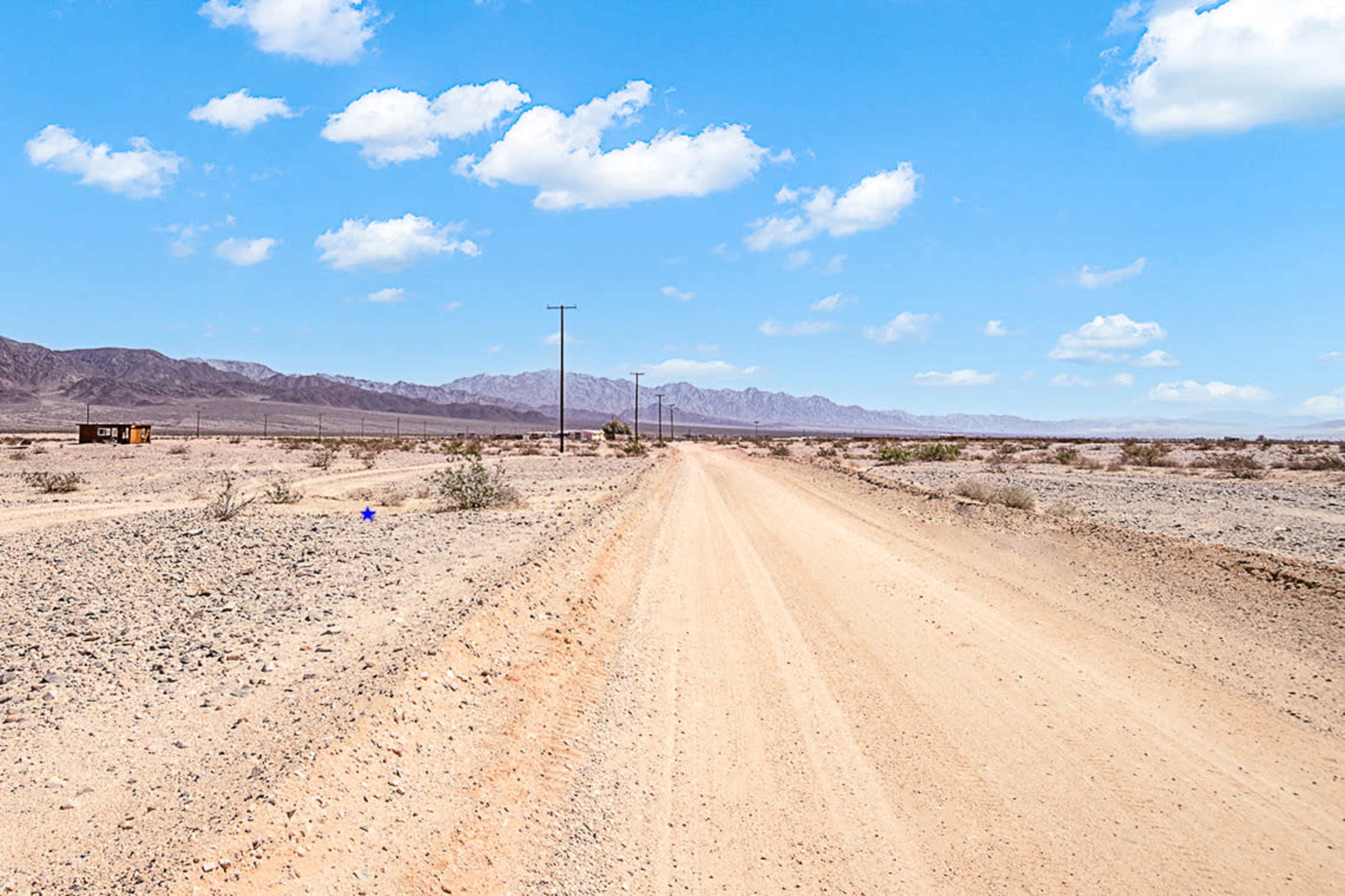 A dirt road stretches through a desert landscape with sparse vegetation and distant mountains under a clear blue sky.