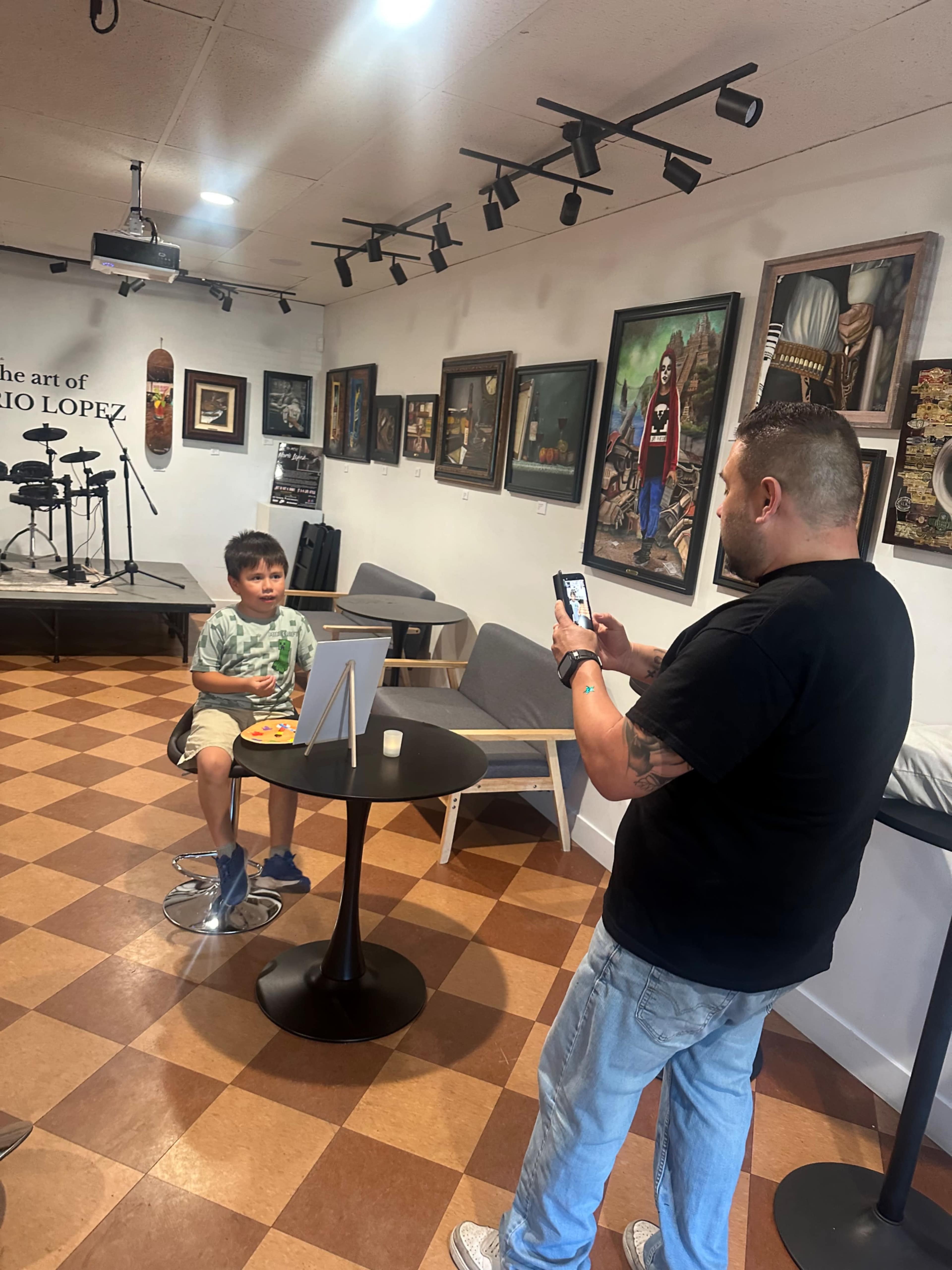 A man is taking a photo of a boy sitting at a table in an art gallery with various paintings on the walls.