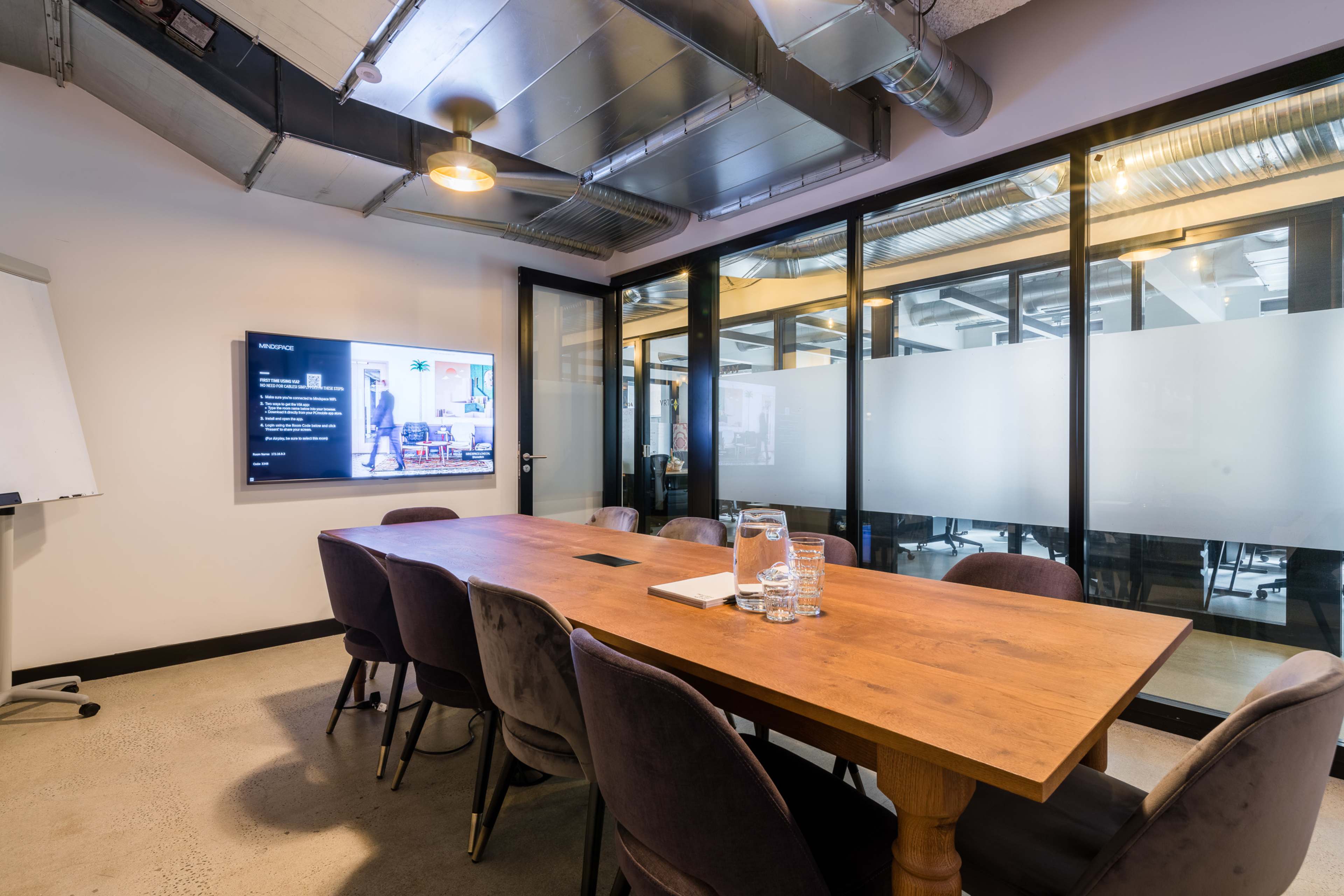 A modern meeting room with a wooden table surrounded by upholstered chairs, a wall-mounted screen, and glass partitions.