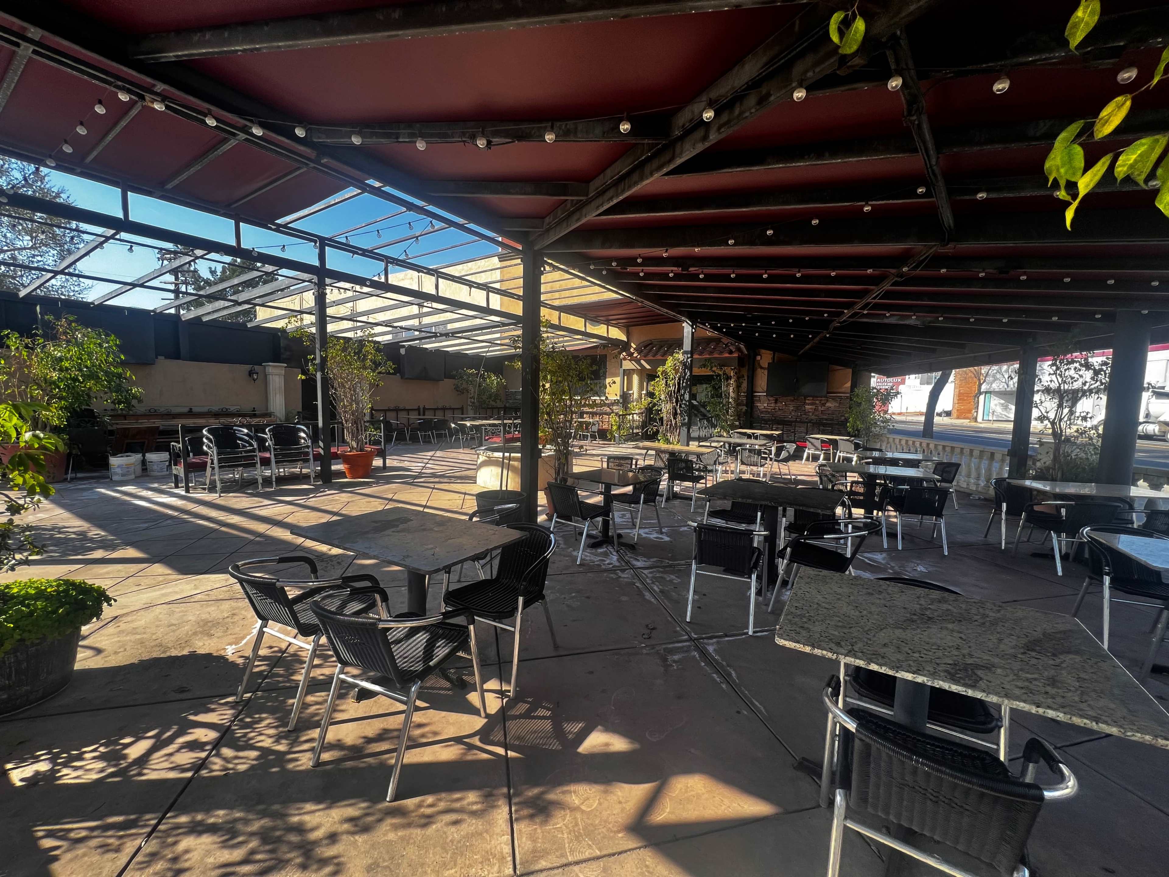 An outdoor dining area with several black metal tables and chairs, surrounded by potted plants and a clear blue sky above.