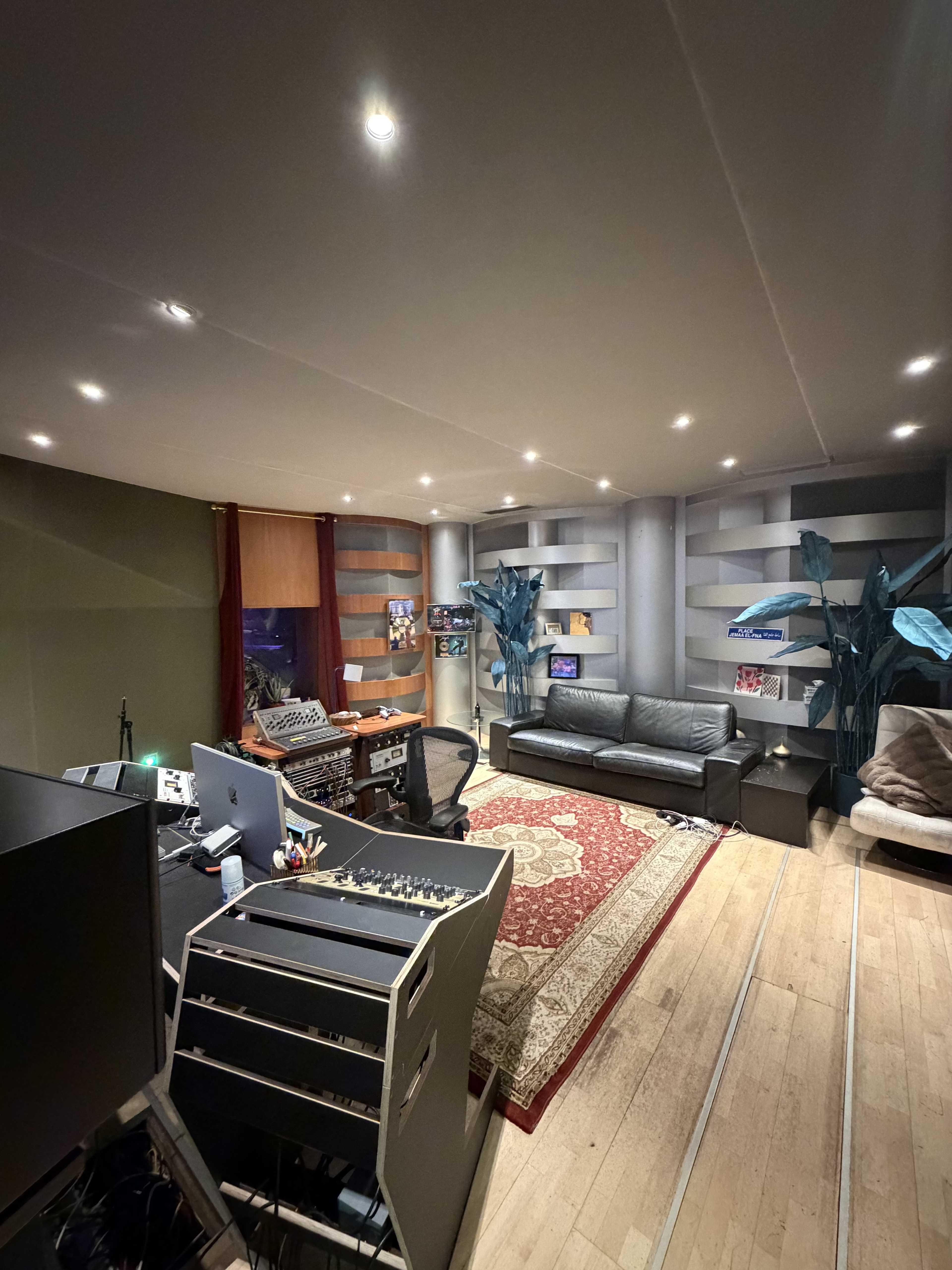 The image shows a music studio featuring a control desk, a couch, various decorative plants, and a rug on the wooden floor.