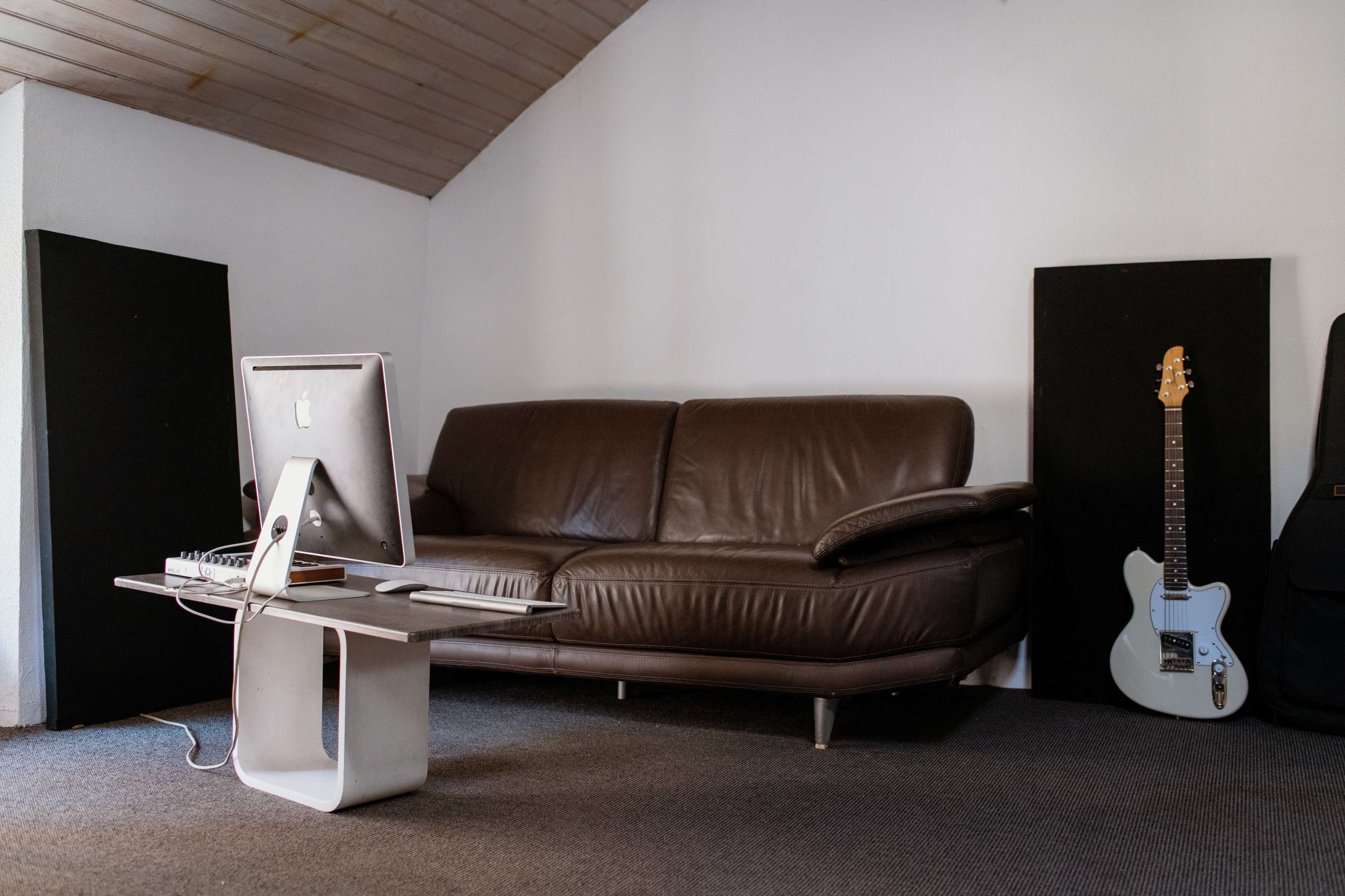 A brown couch is positioned in a room with a computer on a table and a guitar leaning against the wall.