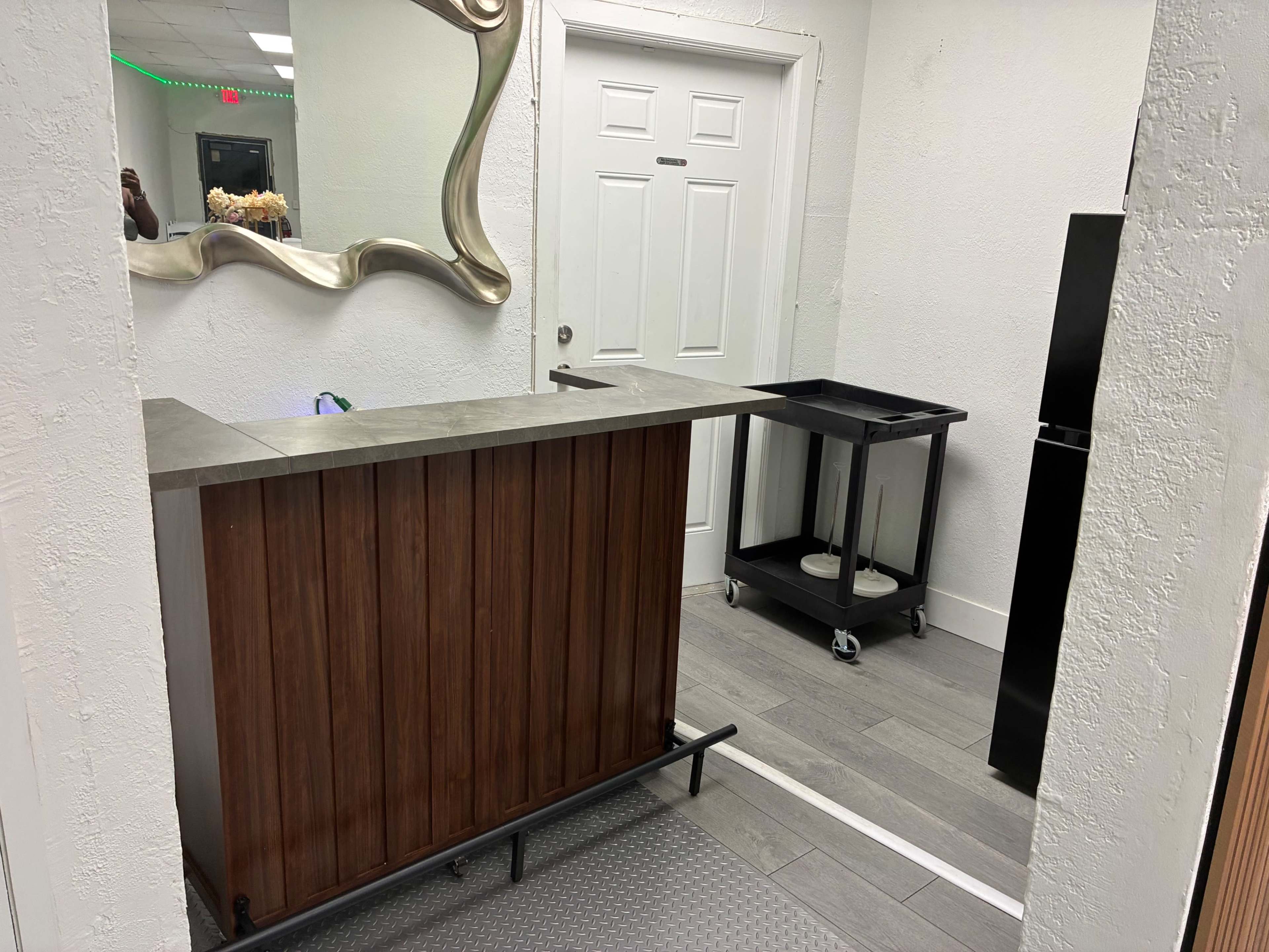 The image shows a small reception area with a wooden counter, a black moveable cart, a door, and a mirror on the wall.