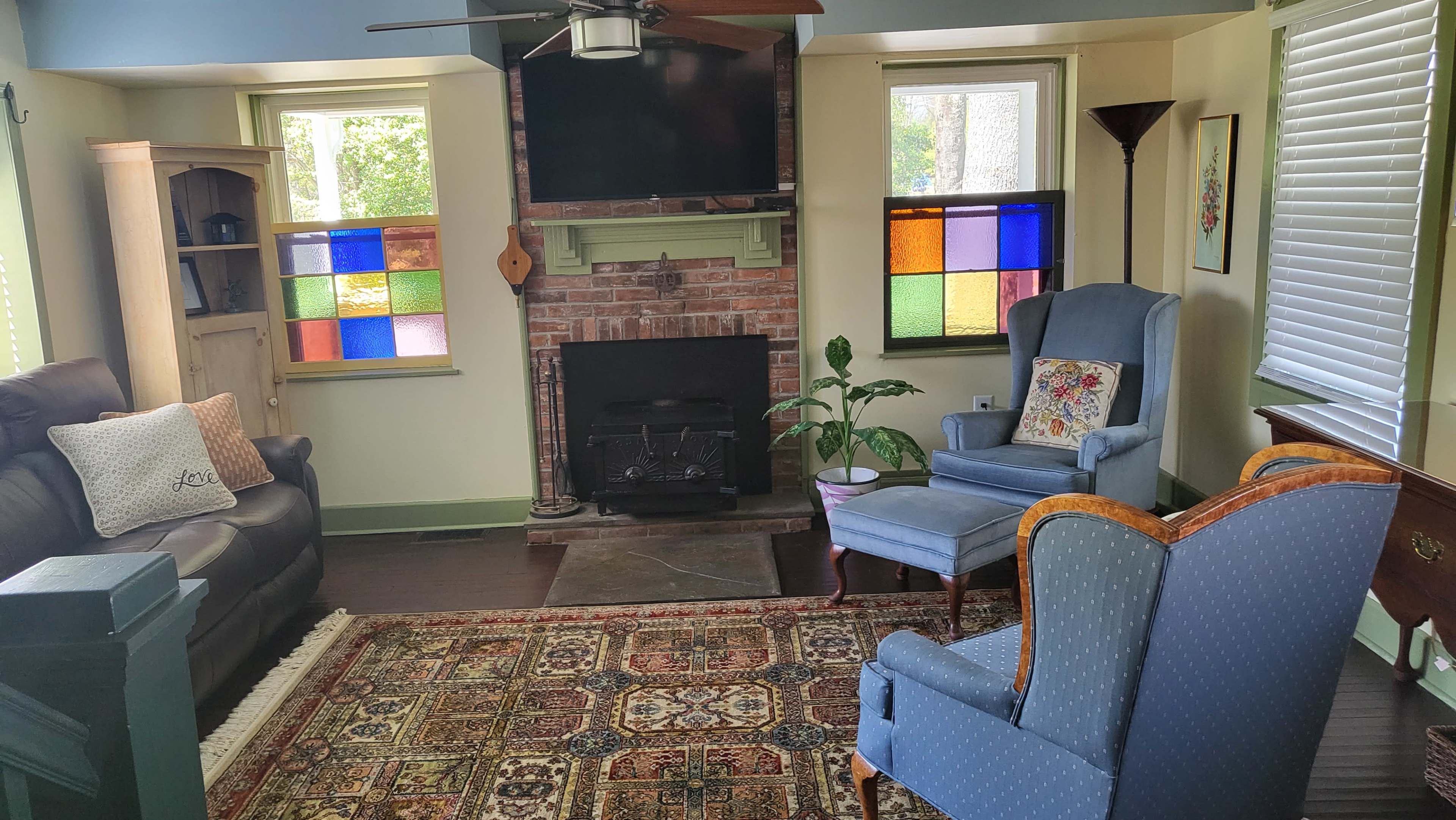 A cozy living room with a brick fireplace, colorful stained glass windows, a television mounted on the wall, and two upholstered armchairs arranged around a patterned rug.