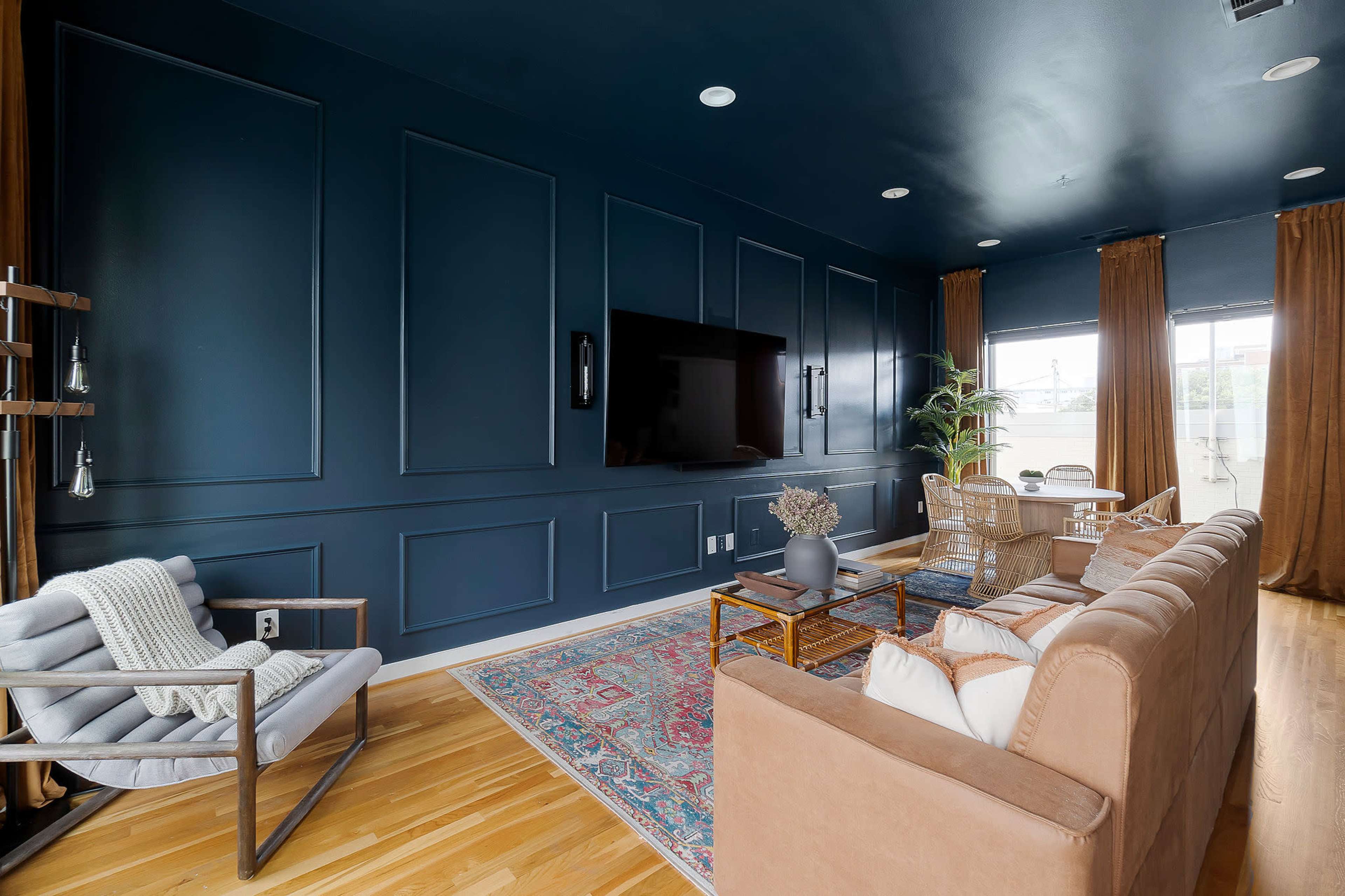 A modern living room featuring dark blue paneled walls, a sofa and armchair, a wooden coffee table, and large windows with curtains.