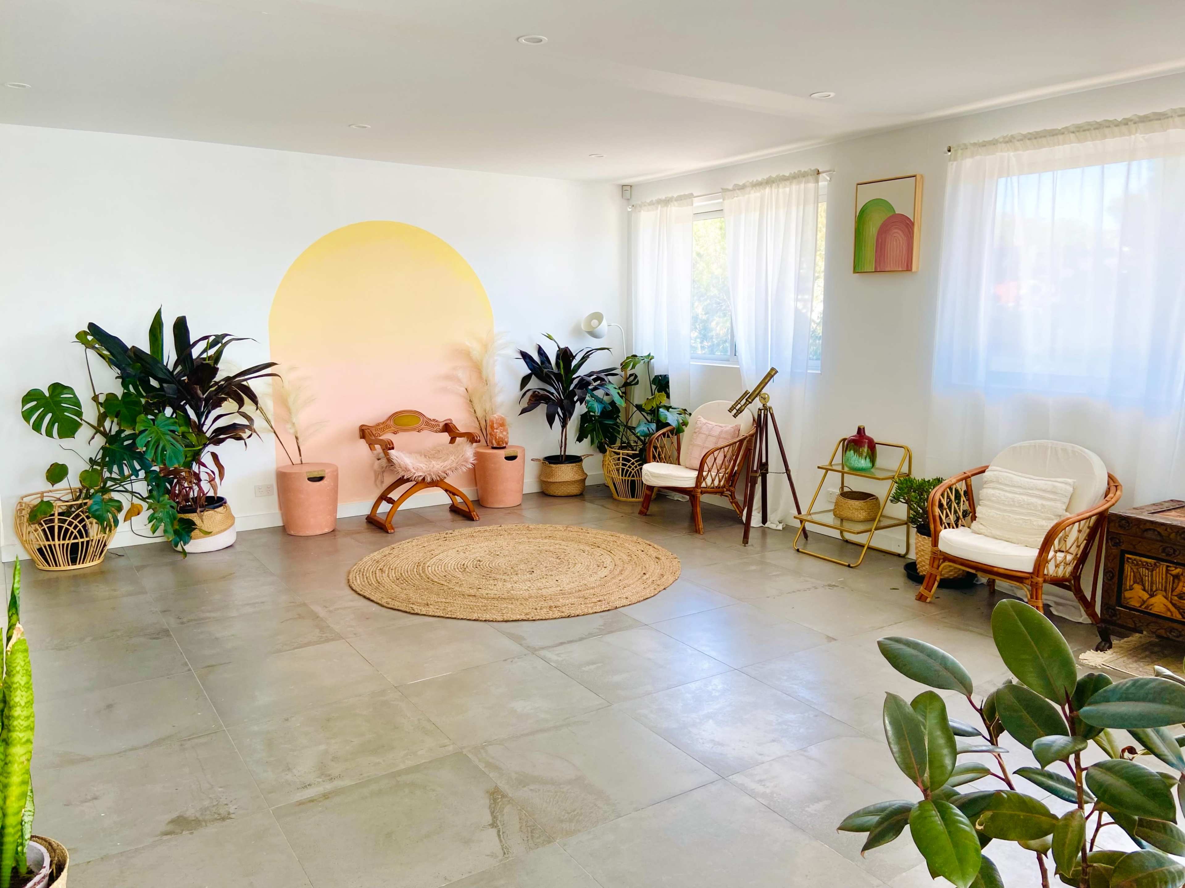 The room features a light, airy design with various plants, a round rug, and wicker furniture against a backdrop of white walls and large windows.