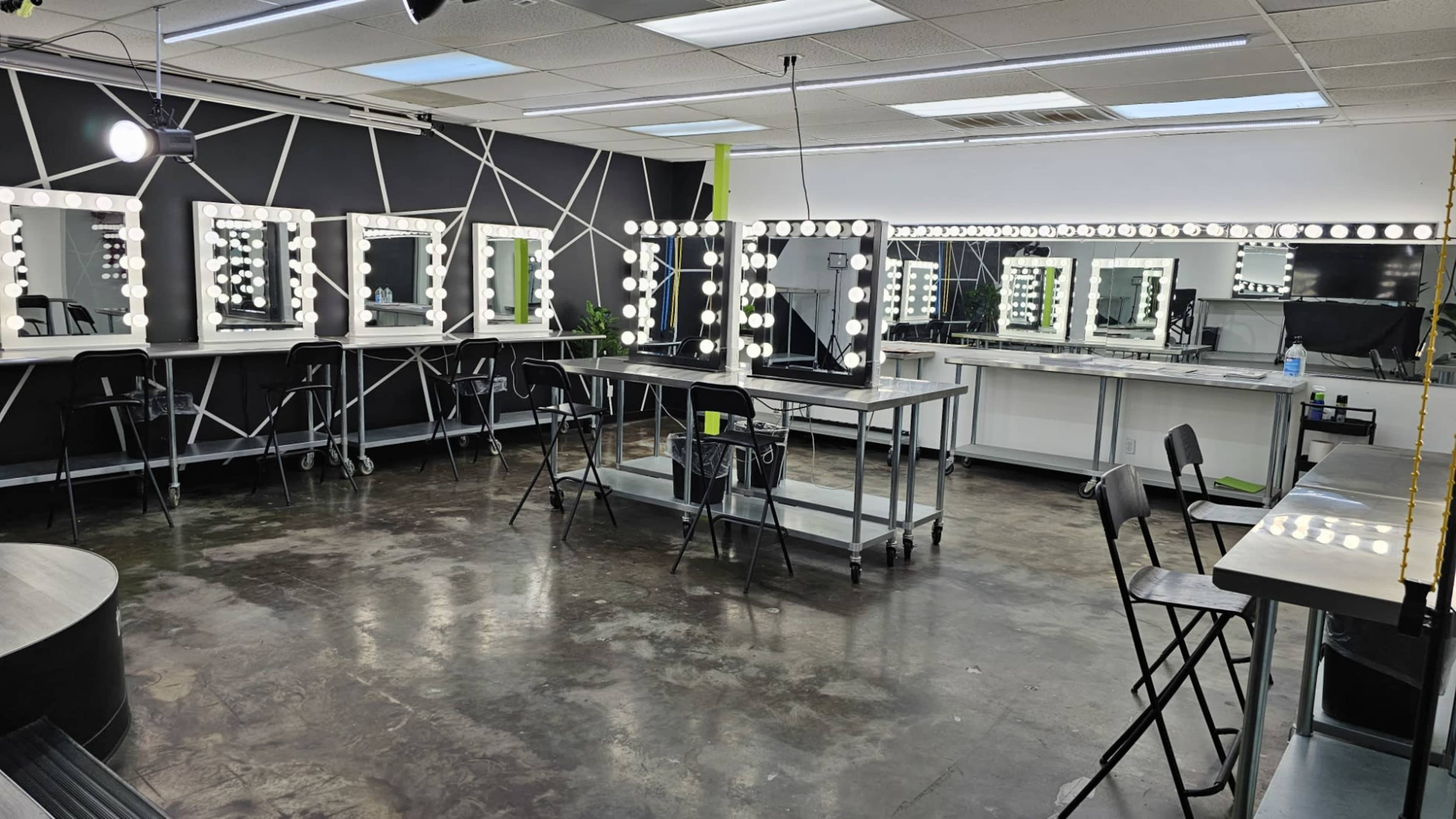 The image shows a spacious makeup studio with multiple illuminated mirrors, chairs, and a central work table.