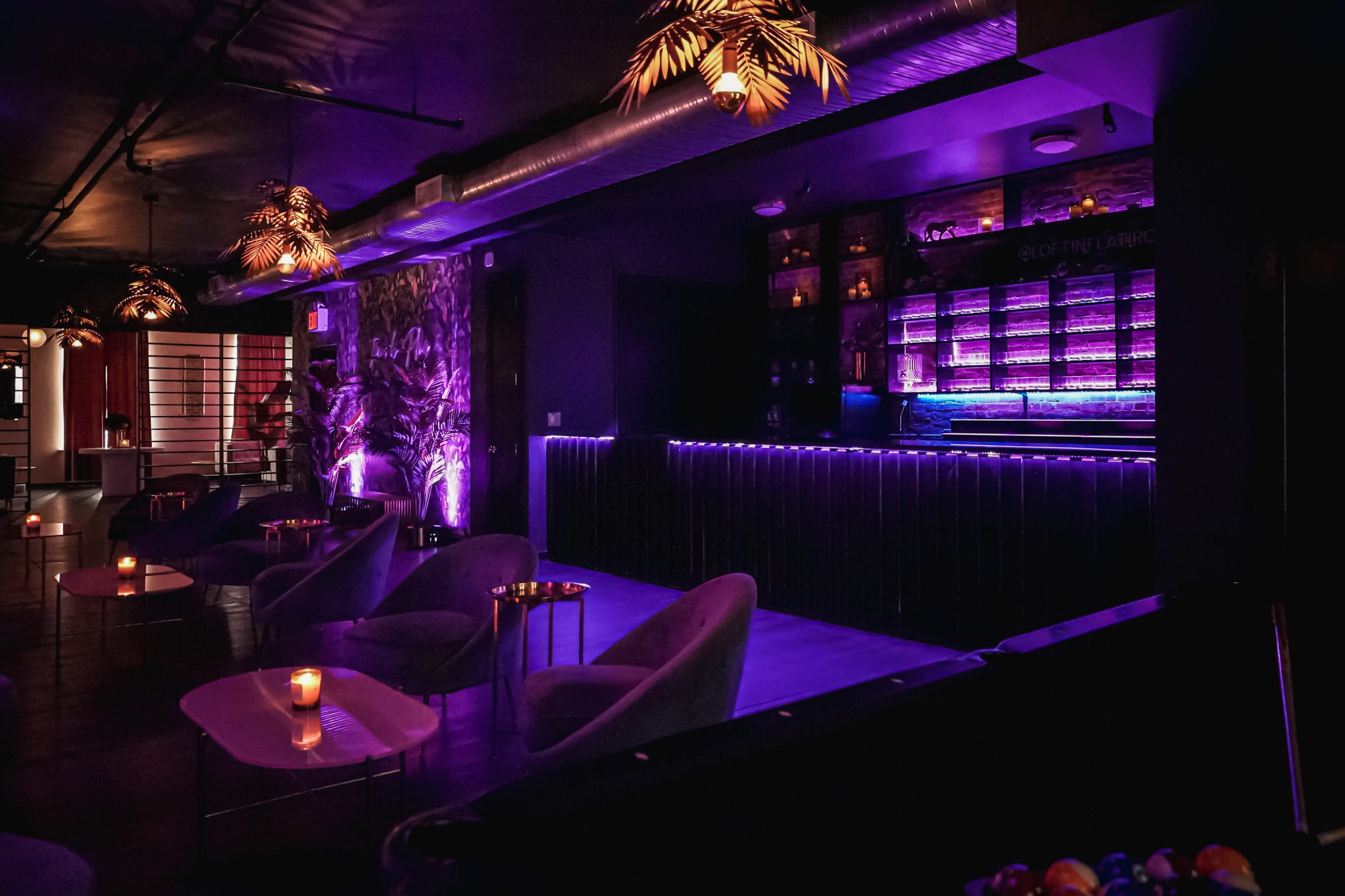 The image shows a dimly lit bar area with purple lighting, featuring modern seating arrangements and a illuminated bar counter.