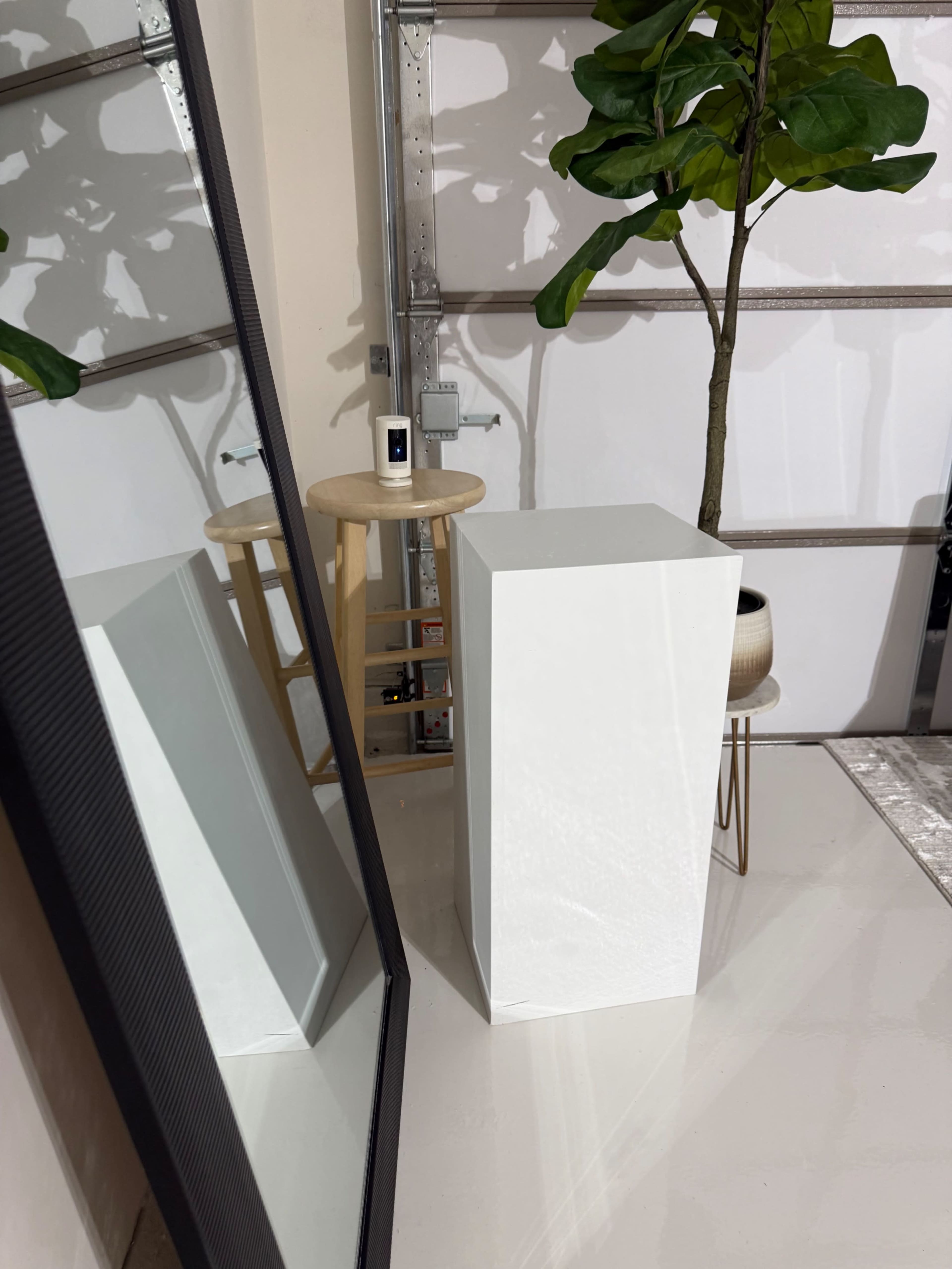 The image shows a minimalist interior scene featuring a white pedestal, a small stool, a potted plant, and a large mirror reflecting these objects.