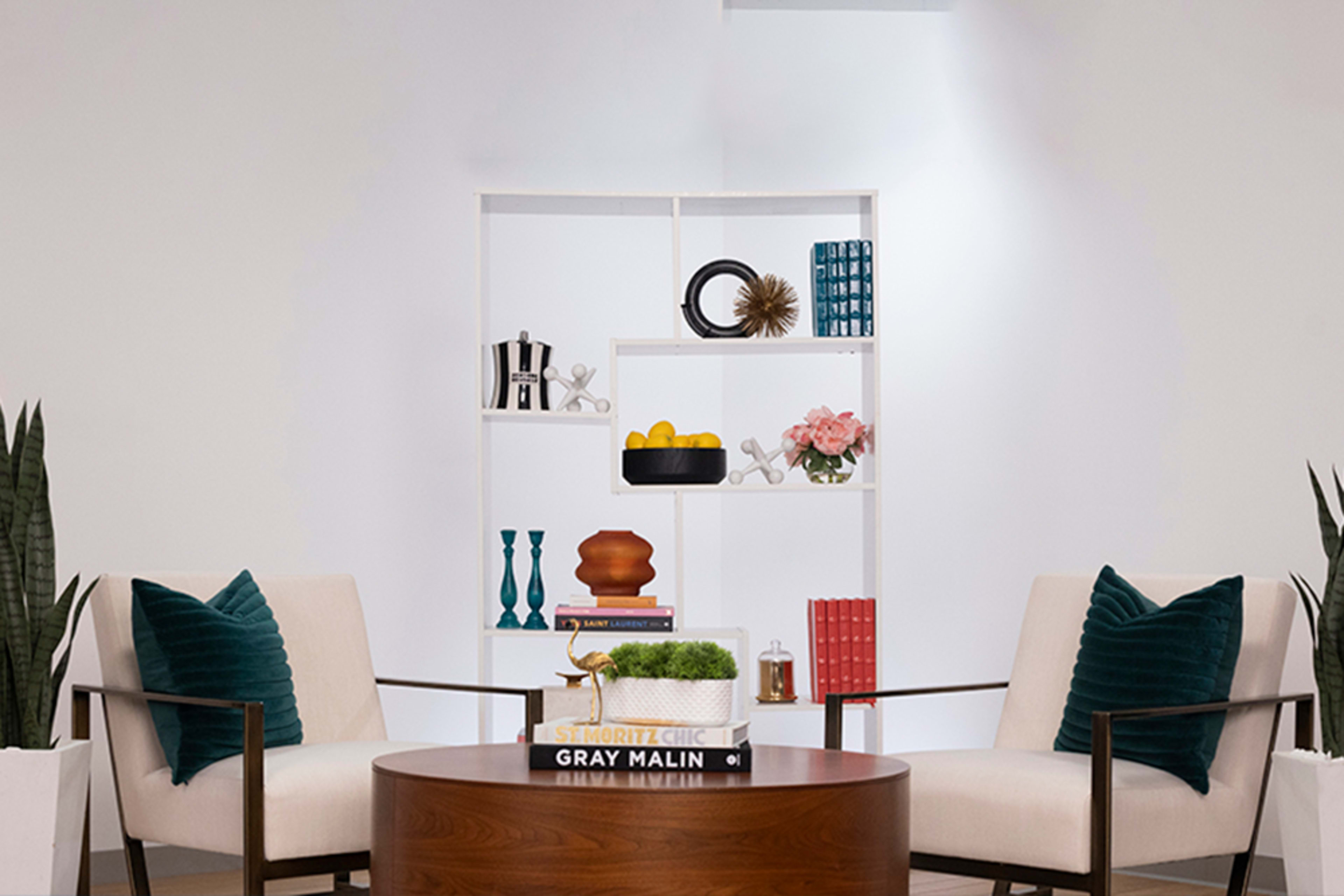 The image features a modern living space with two chairs facing a circular wooden coffee table, while a white shelving unit displays decorative items and plants.