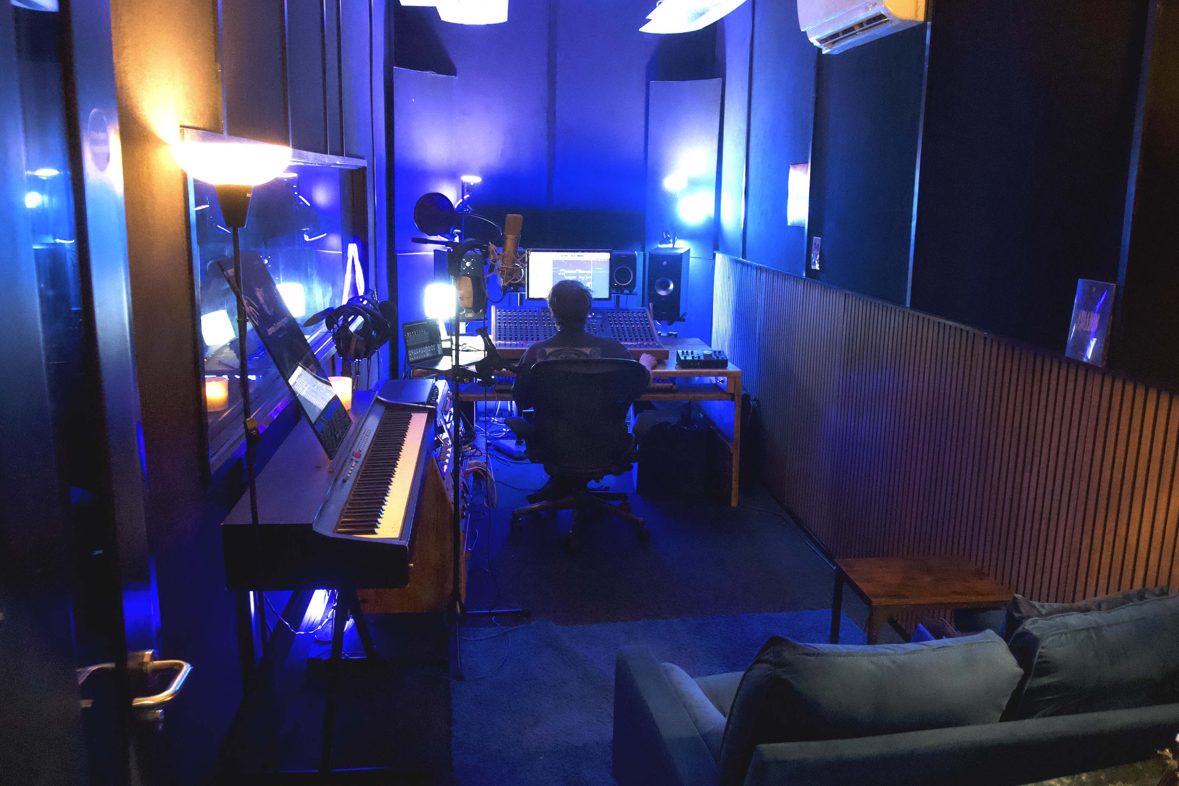 A person sits in a darkened recording studio surrounded by sound equipment, illuminated by blue and white lights.