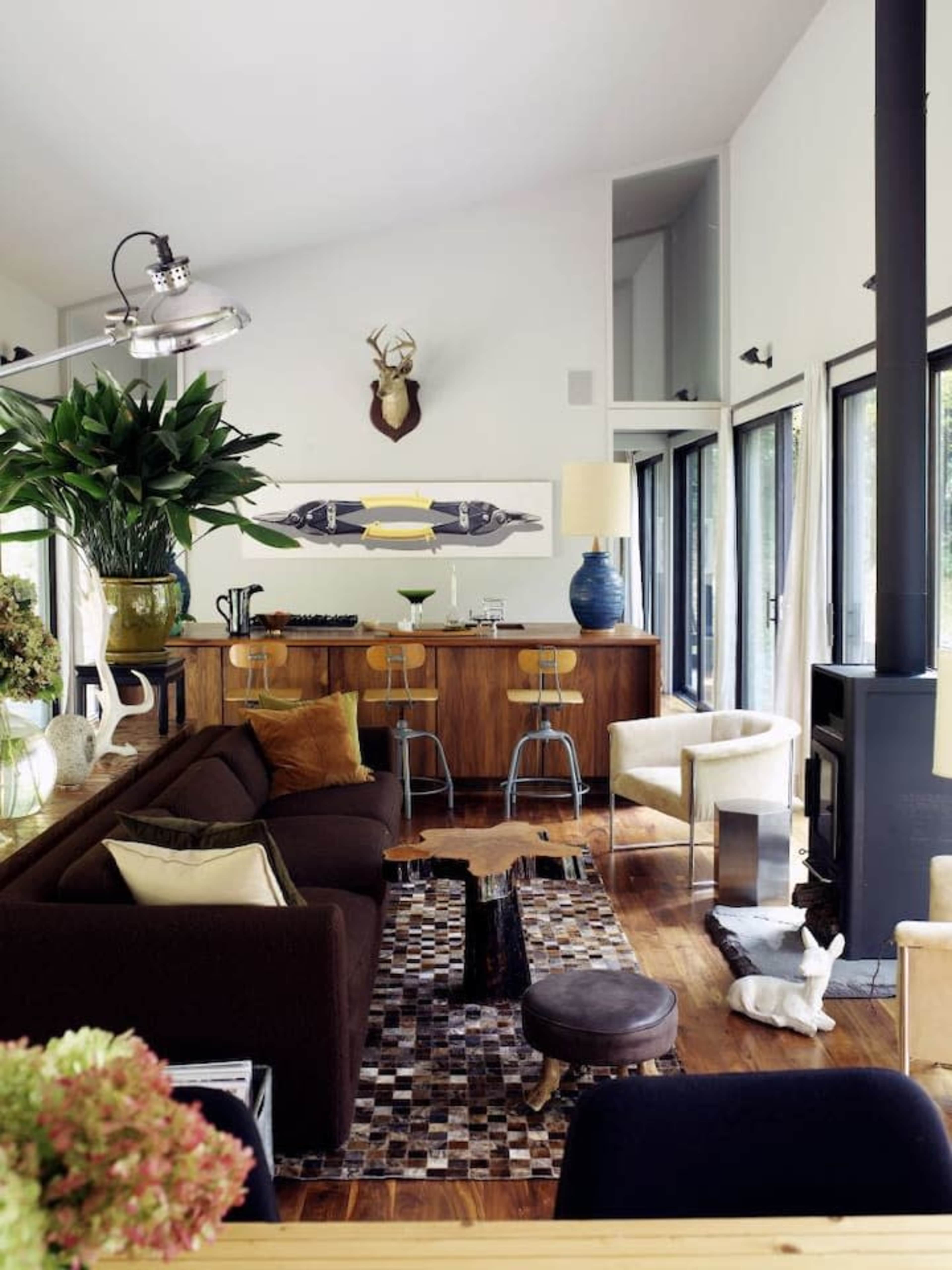 The image shows a modern living room with a dark brown sofa, a patterned rug, and a wooden bar area featuring metallic stools, alongside large windows that provide ample natural light.