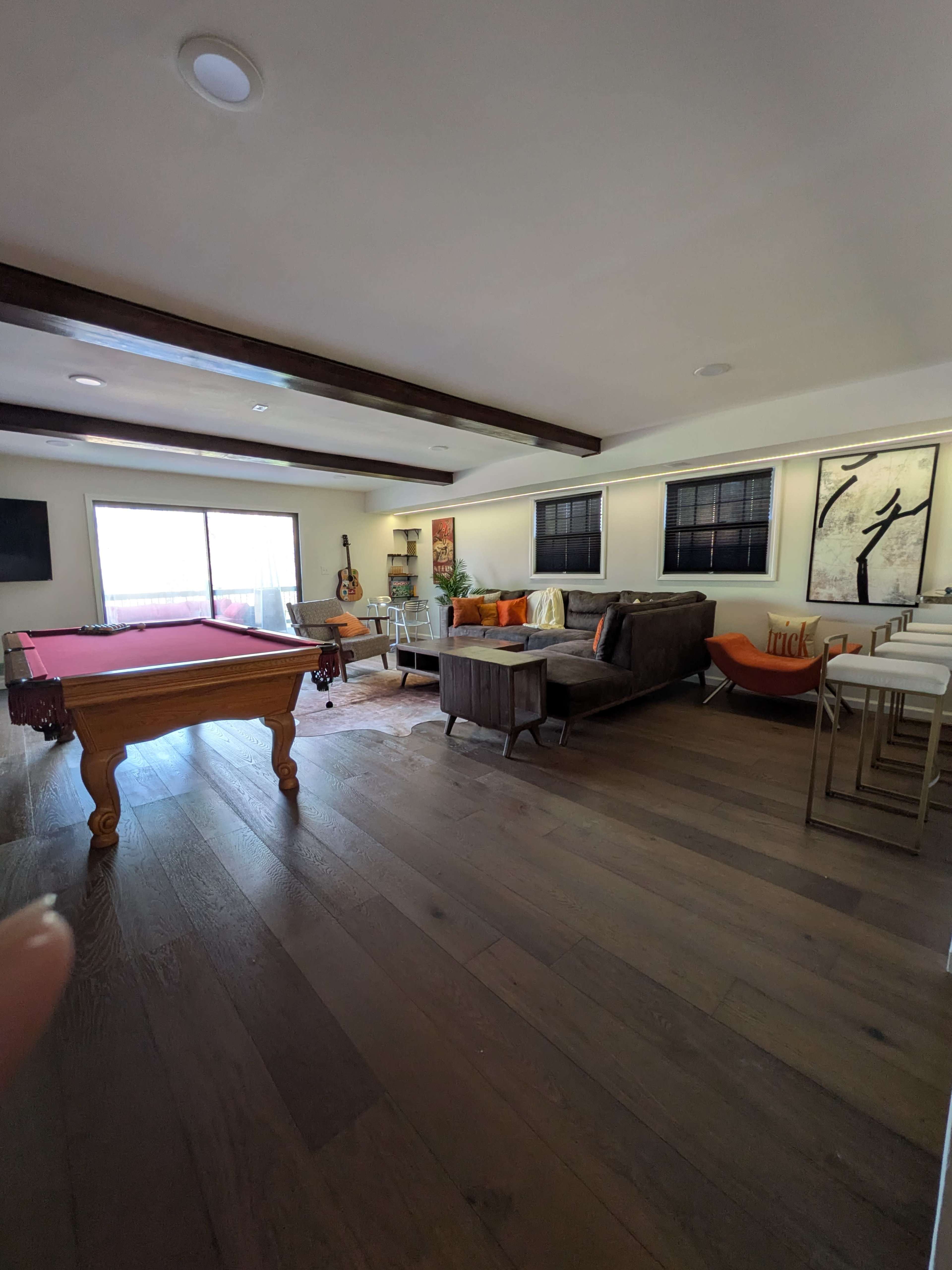 The image shows a spacious living area with a billiard table, a sofa, and a dining table, featuring large windows that allow natural light to enter.