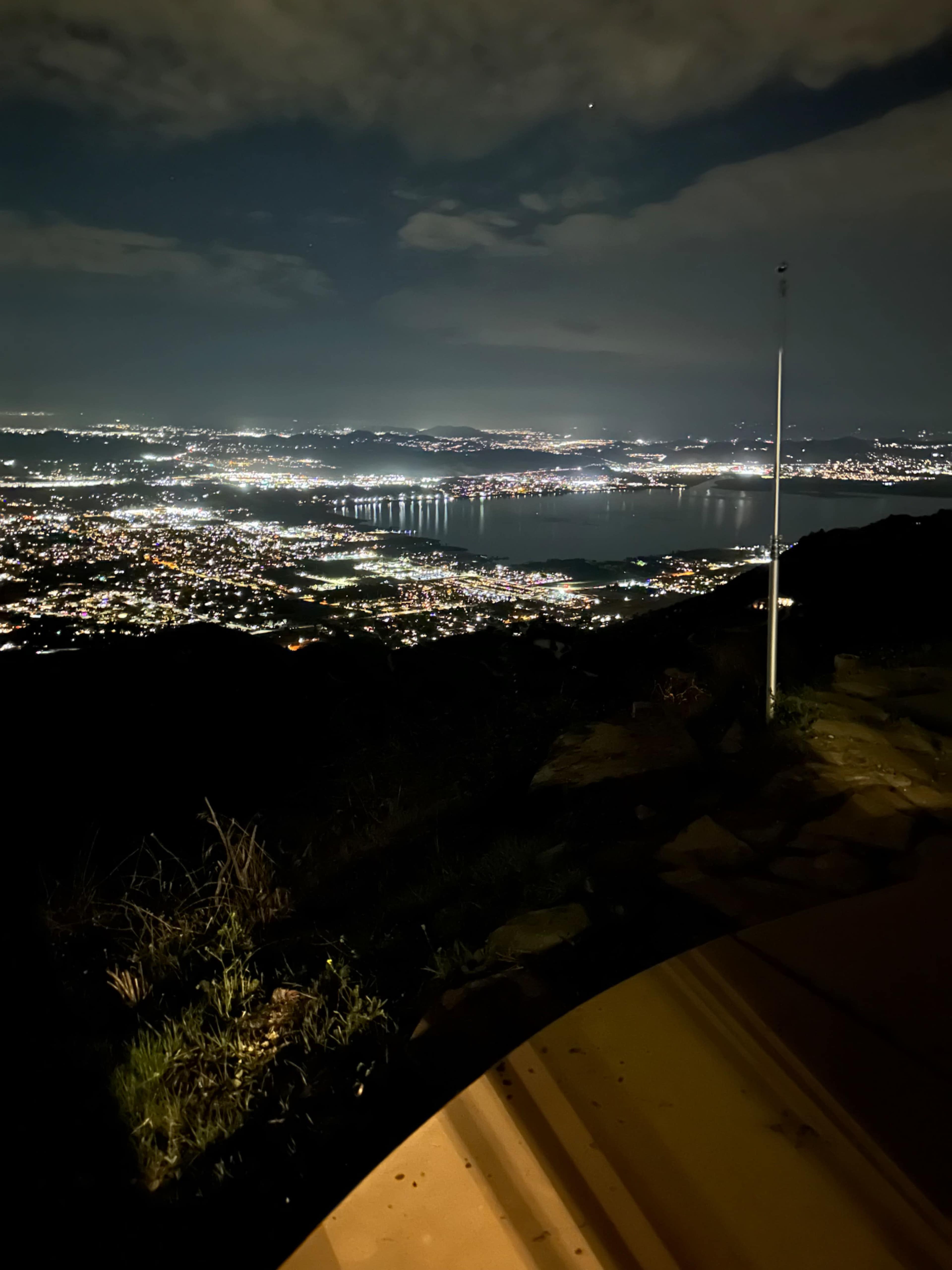 The image shows a nighttime panoramic view of a city landscape illuminated by lights, with a body of water and mountains visible in the distance.