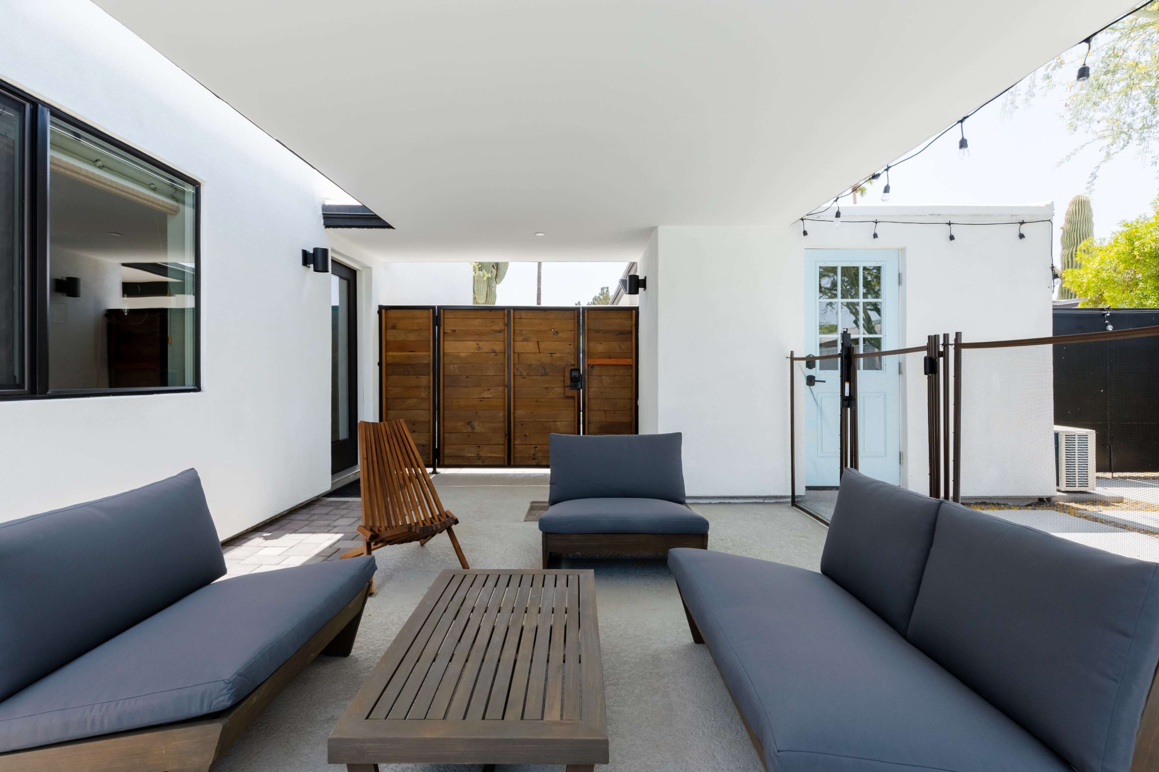 A modern outdoor seating area featuring two gray sofas, a wooden coffee table, and a wooden chair, all situated under a covered patio with a view of a wooden gate.