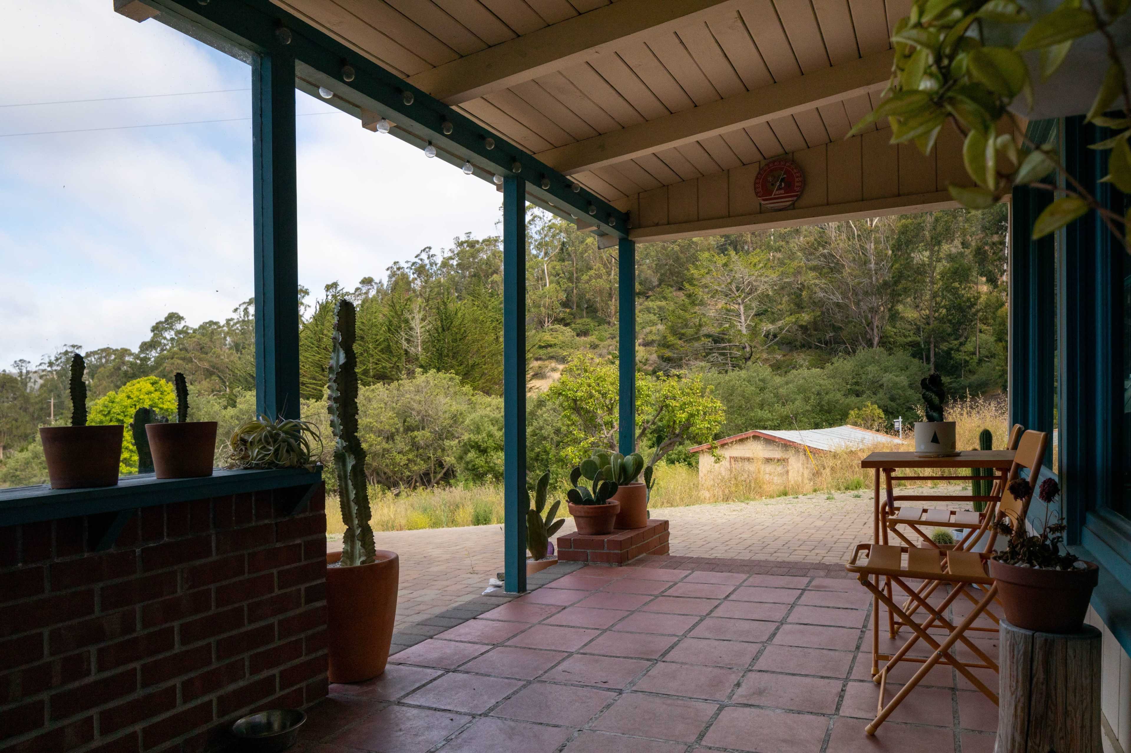 A porch with blue trim features potted cacti and a small wooden table and chair, overlooking a natural landscape.